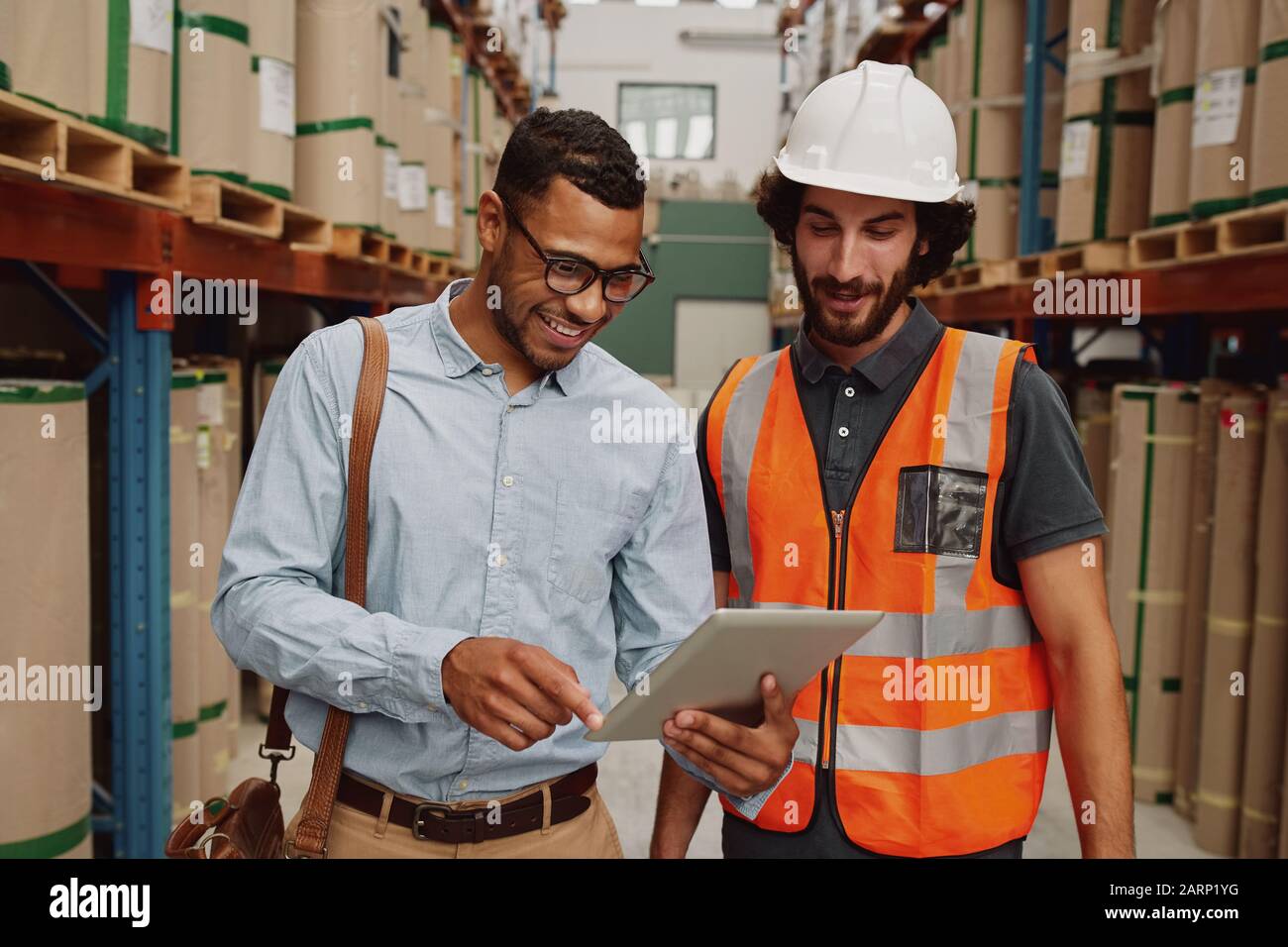Young businessman showing data in touchpad to warehouse worker wearing hardhat and orange jacket uniform Stock Photo