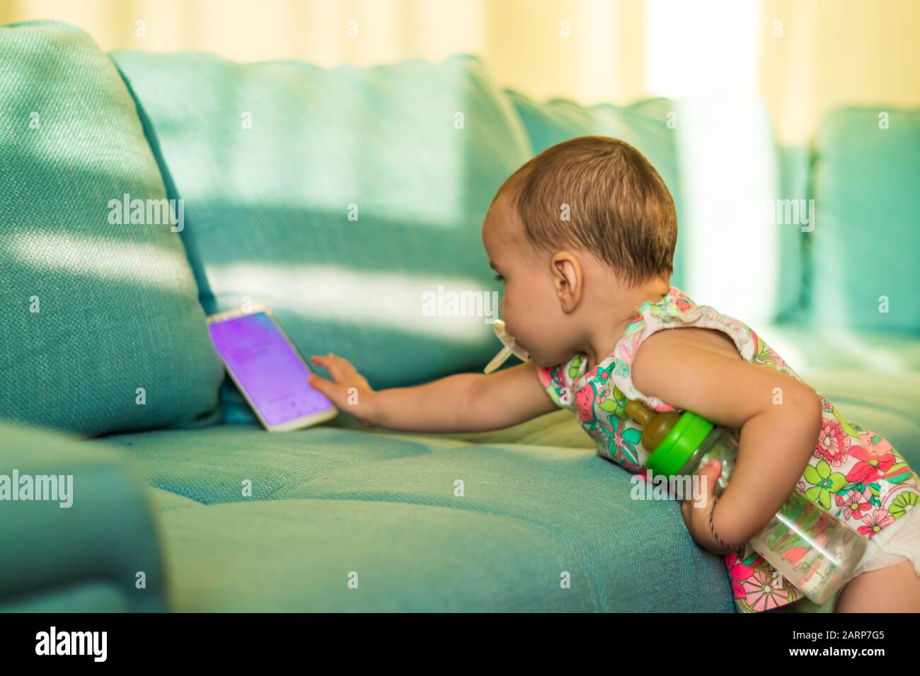 A cute little girl reaching to grab the smartphone from the sofa on a bright sunny day at home. Stock Photo