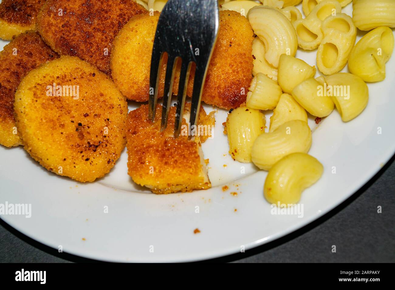 A piece of nuggets on a fork and a white plate with fried chicken nuggets, pasta and ketchup on a dark background. Close up Stock Photo