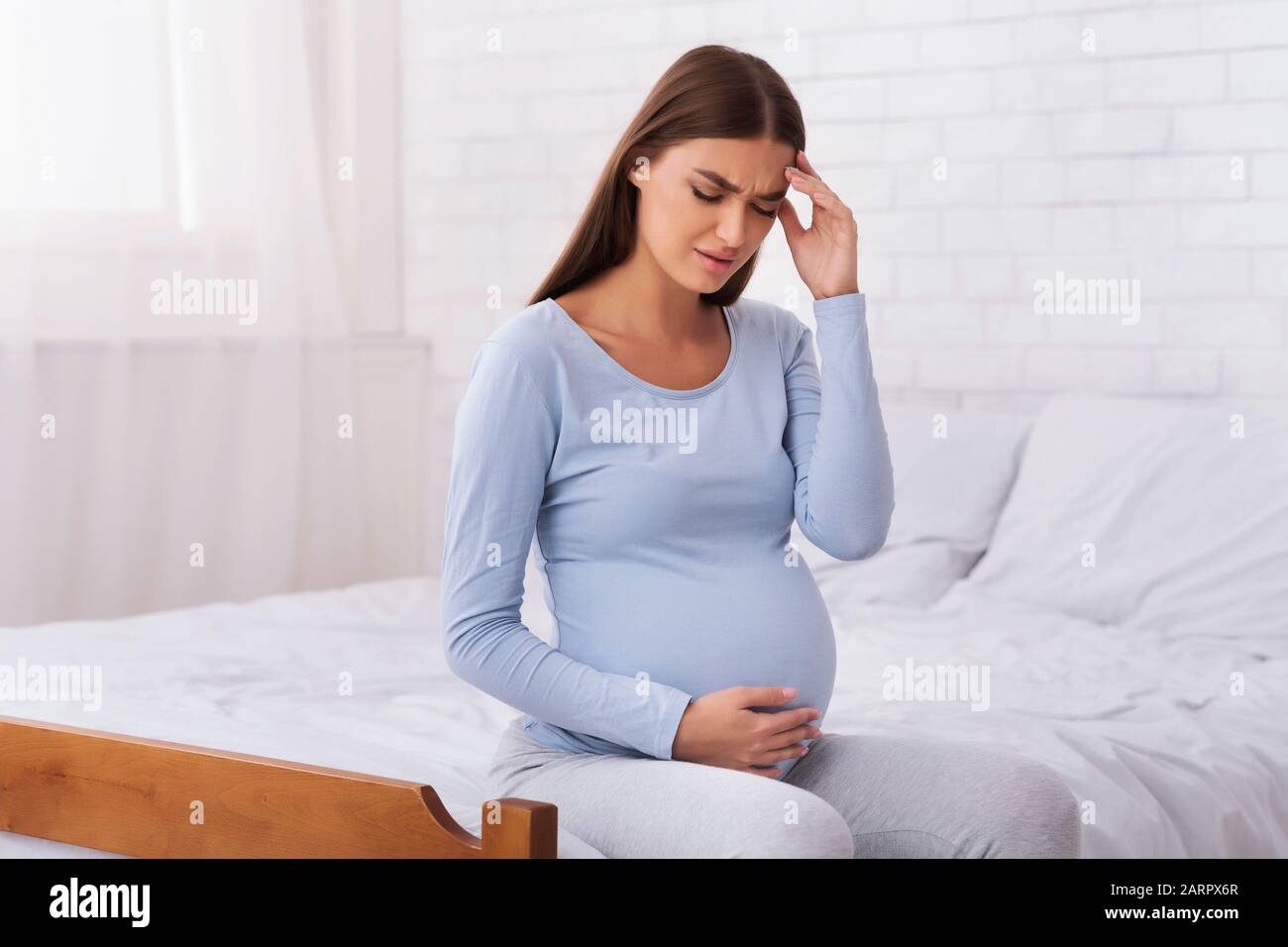 Unhappy Expectant Girl Touching Head And Belly Sitting On Bed Stock Photo