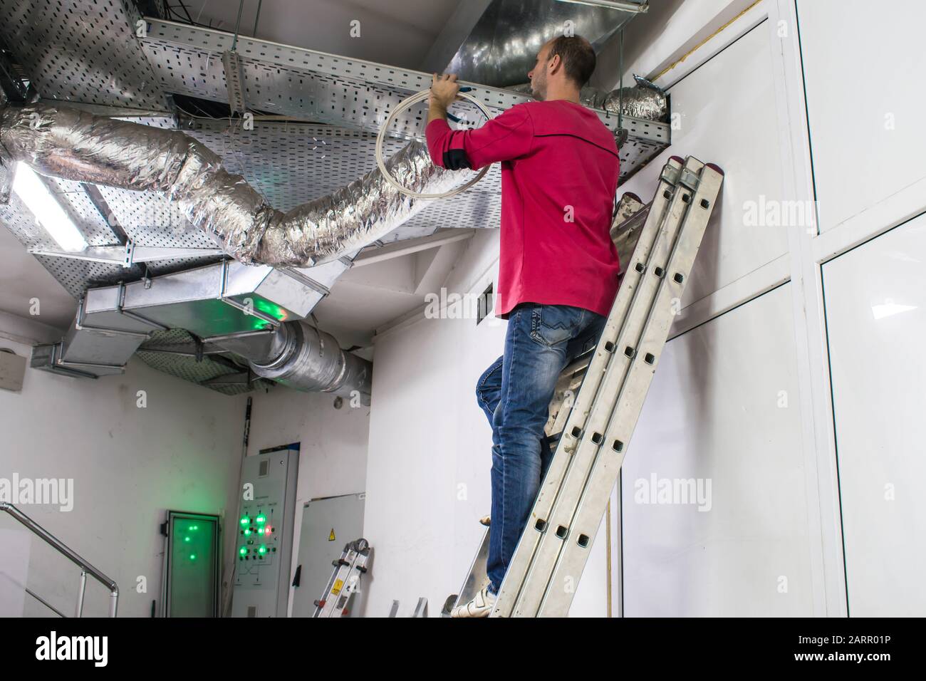 A man standing on the stairs is repairing the ventilation ducts. Maintenance of communications is performed in the technical room. The installer works Stock Photo