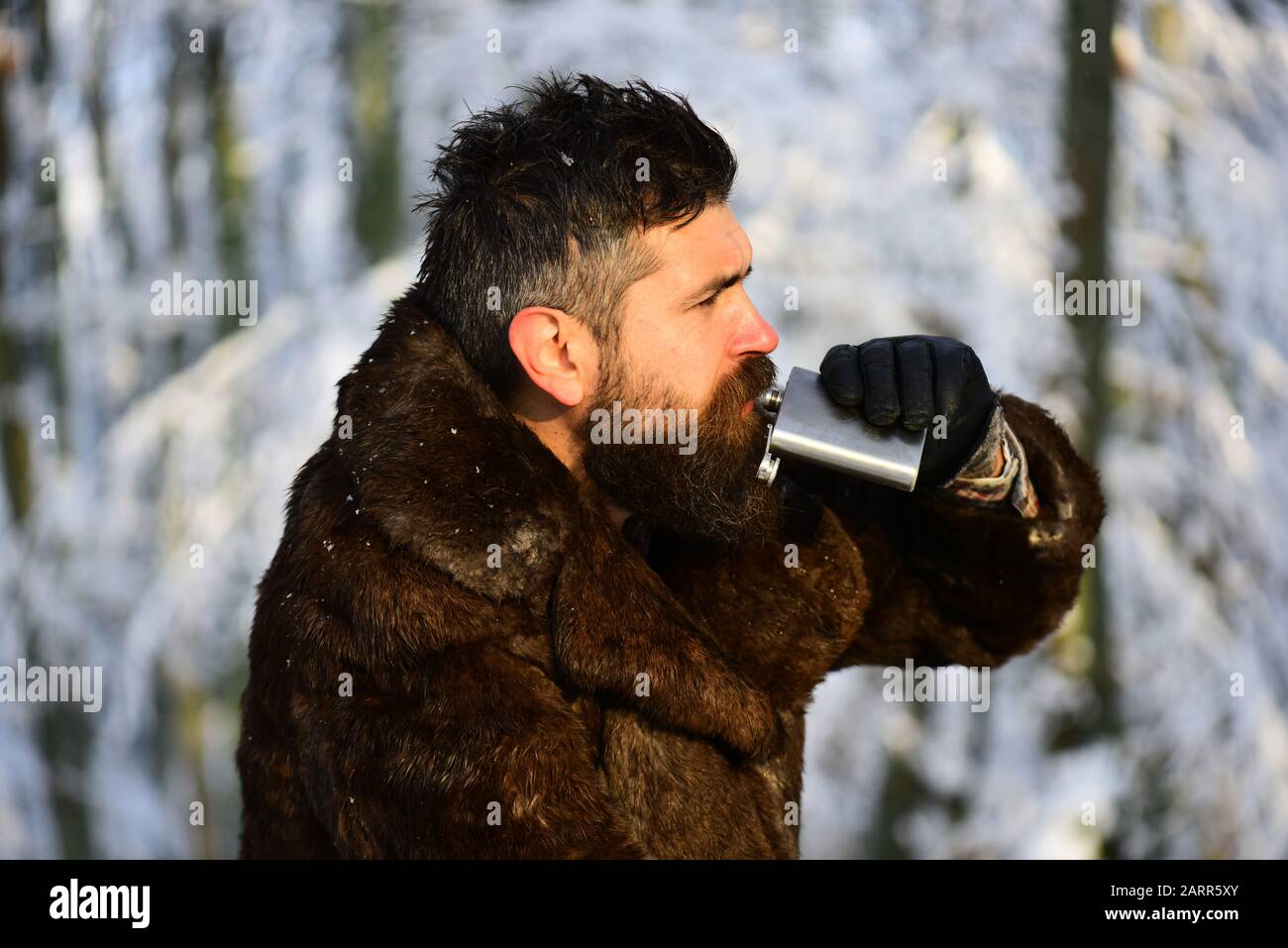 Macho with beard and mustache in forest. Man in fur coat drinks from metal flask. Warming drinks concept. Guy on snowy nature park background, defocused. Stock Photo
