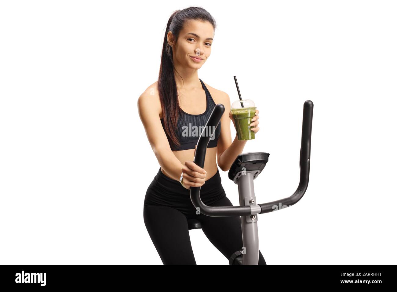 Young woman riding a stationary bike and holding a green healthy smoothie isolated on white background Stock Photo