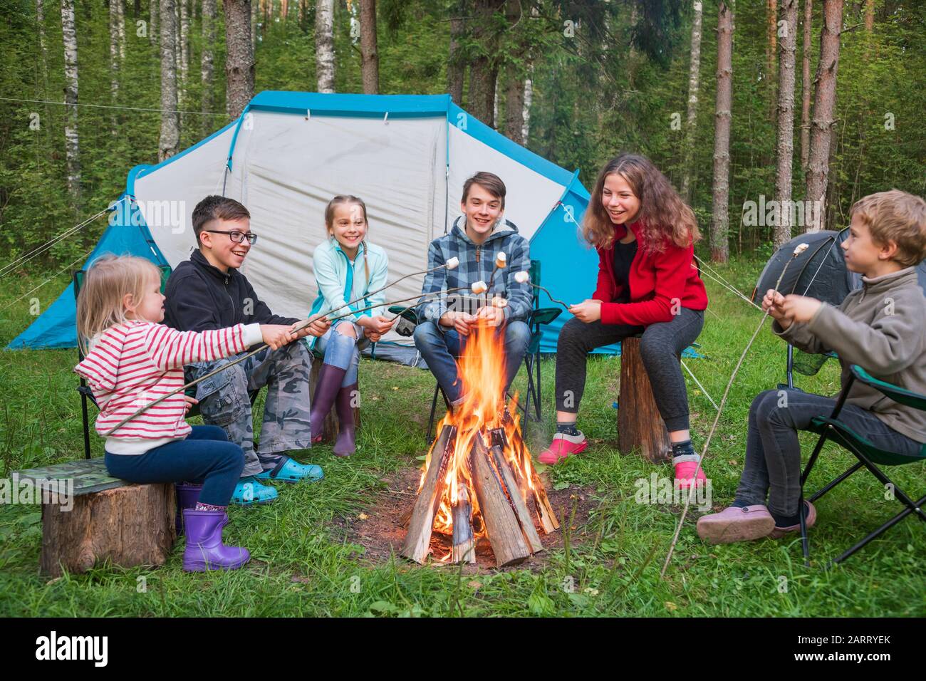 Six children of different age sitting together around burning bonfire having fun roasting marshmallows while camping in a summer forest. Camping tents Stock Photo