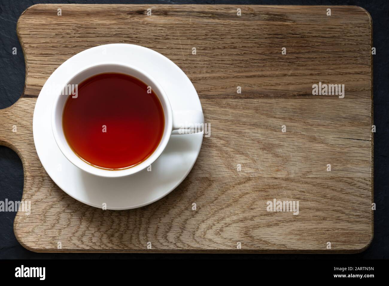 Overhead shot of clear Rooibos tea in white cup and saucer on a wooden chopping board with black slate below. Stock Photo