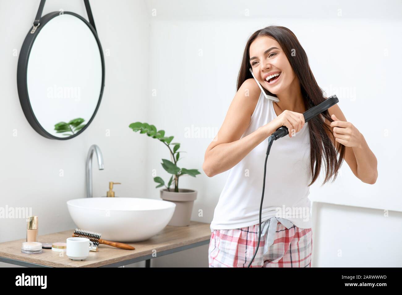Morning of beautiful young woman straightening hair while drinking coffee  and talking by phone in bathroom Stock Photo - Alamy