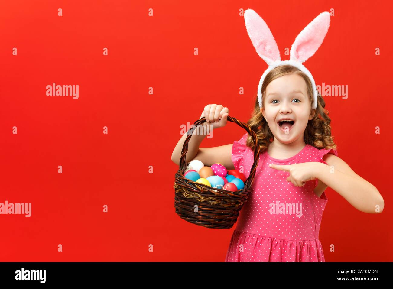 Enthusiastic child in the ears of a bunny on a red background. Little girl shows a finger on a basket with Easter eggs. Stock Photo
