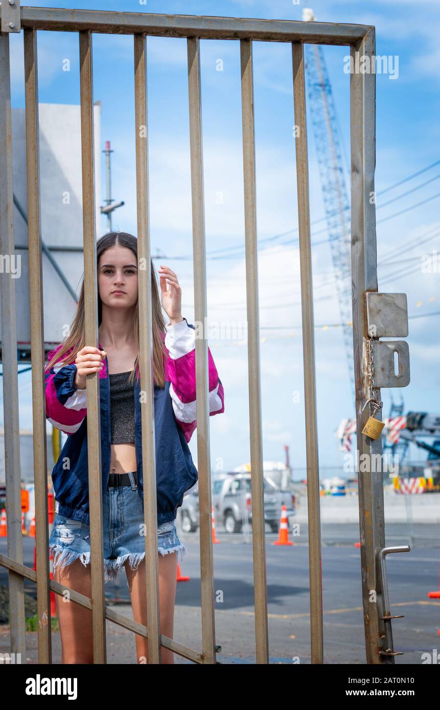 Teenager girl behind bars of a gate contrasts with harsh industrial setting background. Stock Photo