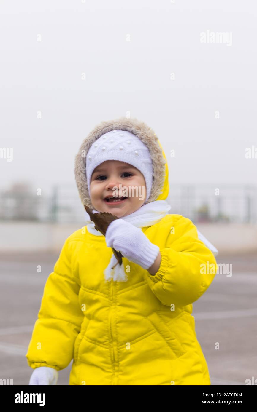 A cute girl in a yellow winter jacket holding a big brown leaf in front of her face outdoor in a foggy day. Stock Photo