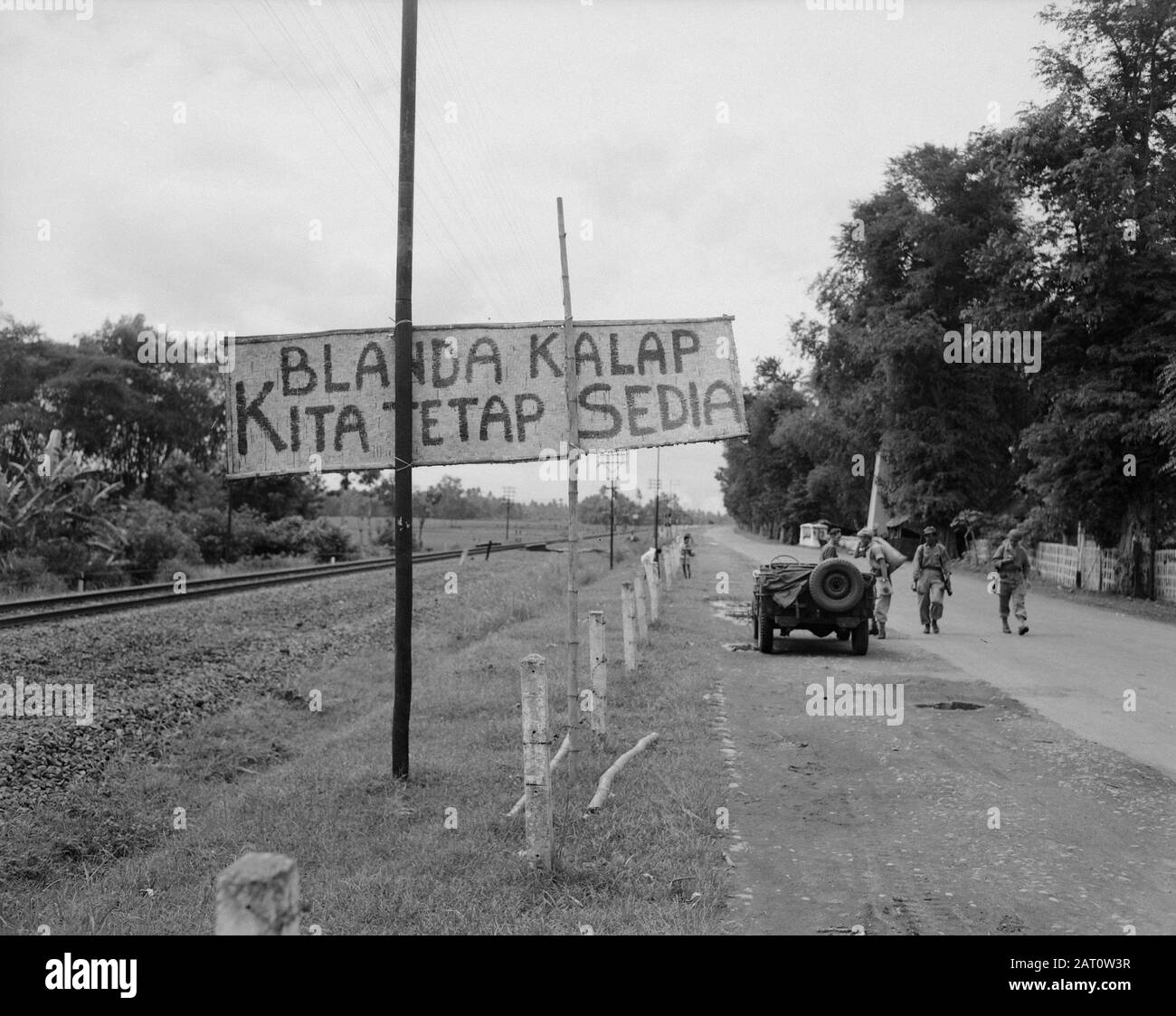 Army Information Service Surabaya An army jeep stands along a long road and  a railway line. There are four soldiers. [on a plate the text 'Blanda kalap  kita tetap sedia' Annotation: Translated.