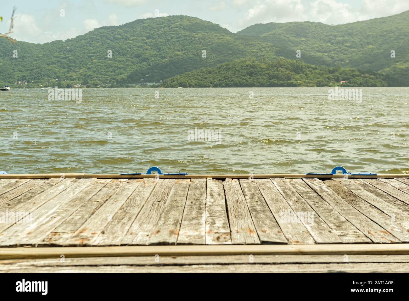 Old wooden pier over a lake (Lagoa da Conceição - Florianopolis - Brazil). Lake with a ripples surrounded by vegetation and mountains on a sunny day. Stock Photo