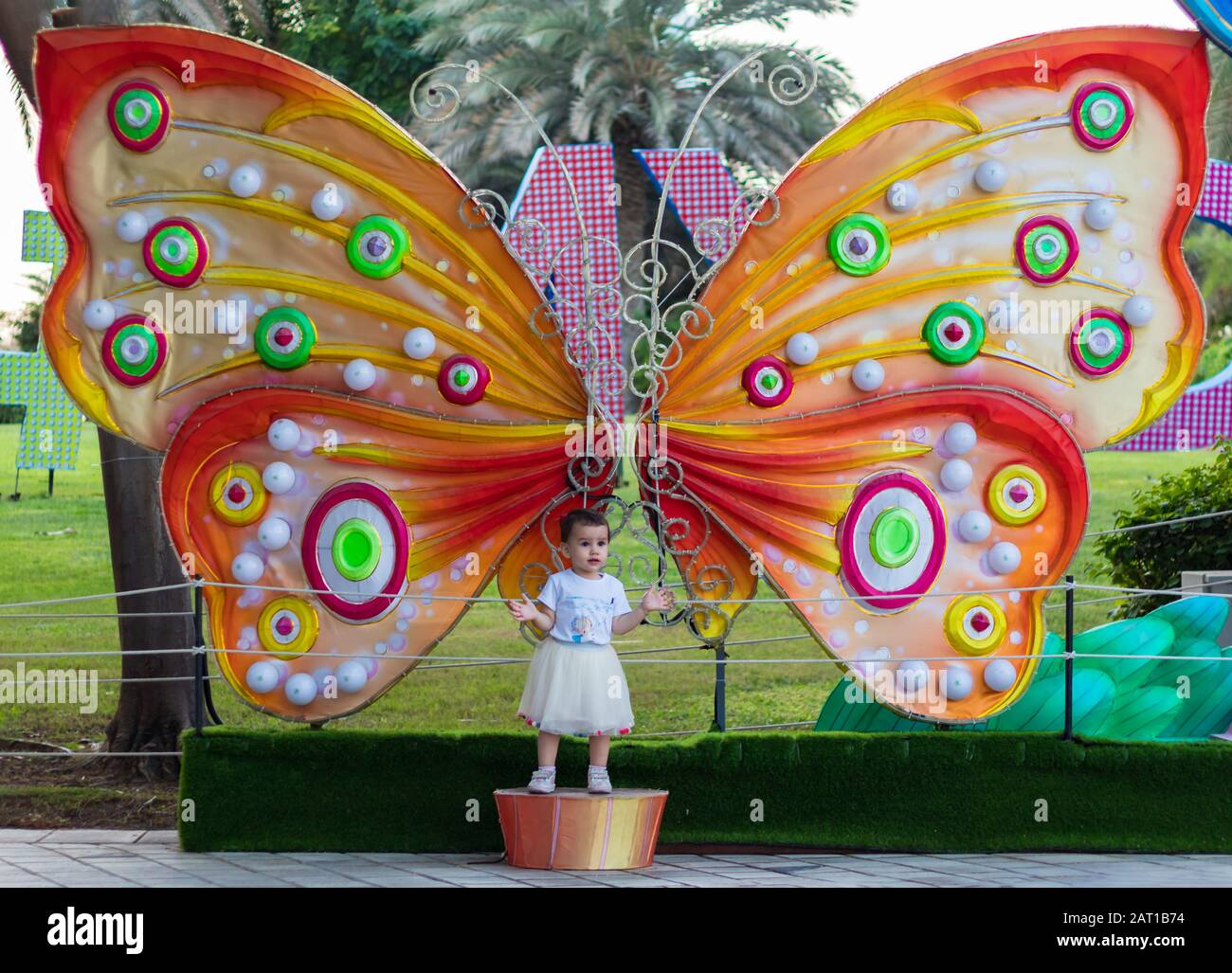 a cute little girl in a white dress posing in front of the huge butterfly in the Dubai Glow Garden. Stock Photo