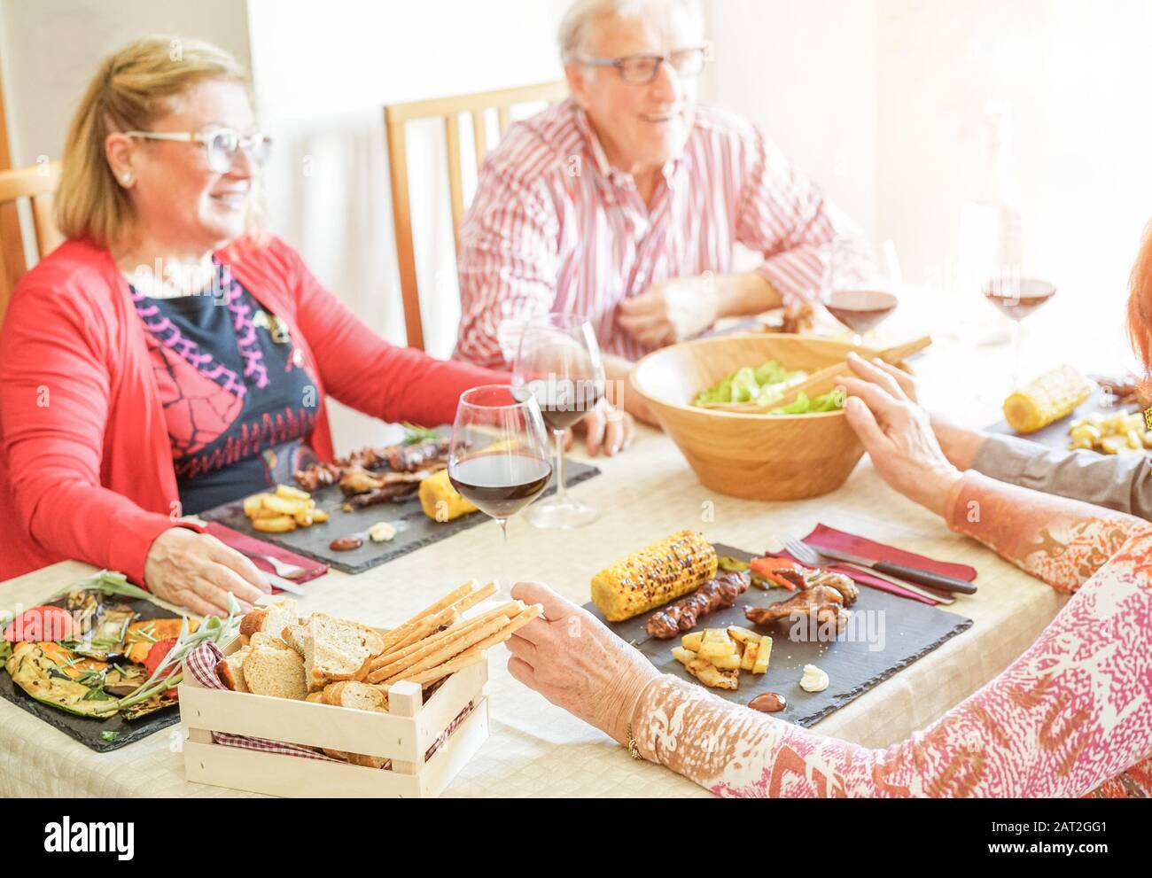 Senior happy friends having lunch meal indoor - Mature people laughing and eating bbq meat and vegetables at home - Joyful elderly lifestyle concept - Stock Photo