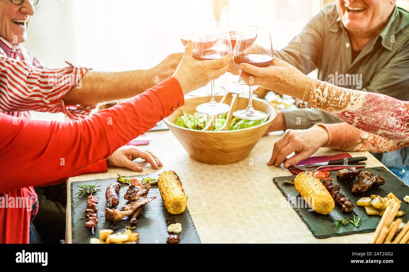 Senior friends cheering with red wine at barbecue lunch indoor - Old people laughing and eating bbq meat with vegetables, corn cobs and salad - Focus Stock Photo