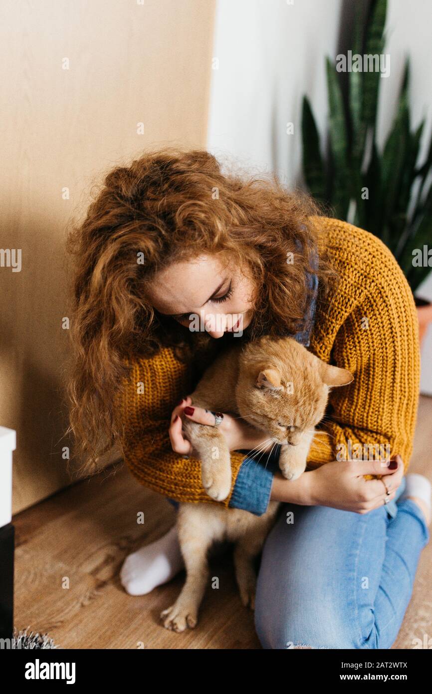 Portrait of beautiful woman with curly hair playing with yellow cat indoors, in house. Stock Photo