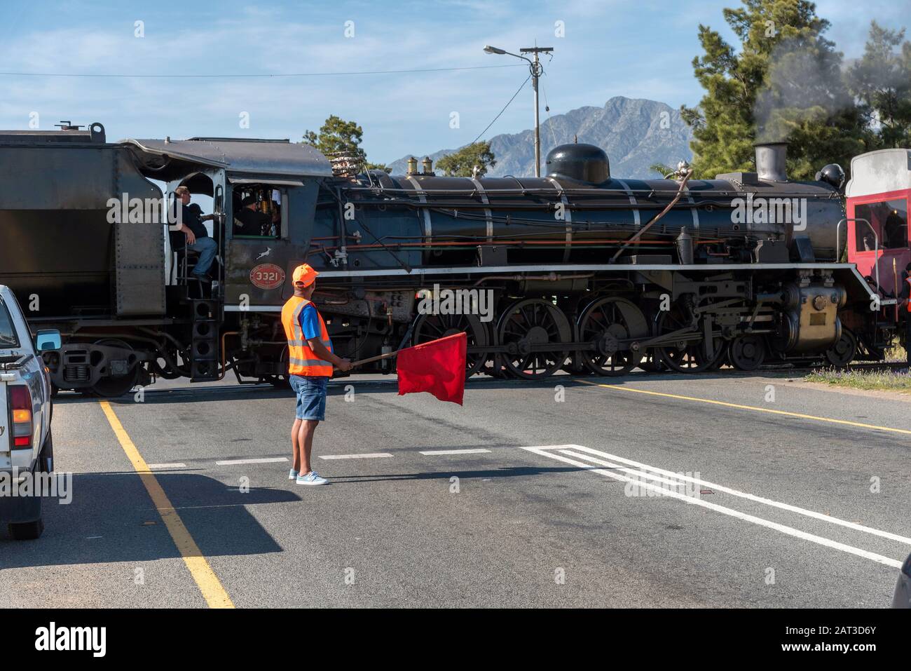 Wolseley, Swartland region, South Africa. 2019. Man with red flag holds traffic. Steam train diesel powered crossing highway at Wolseley. South Africa Stock Photo