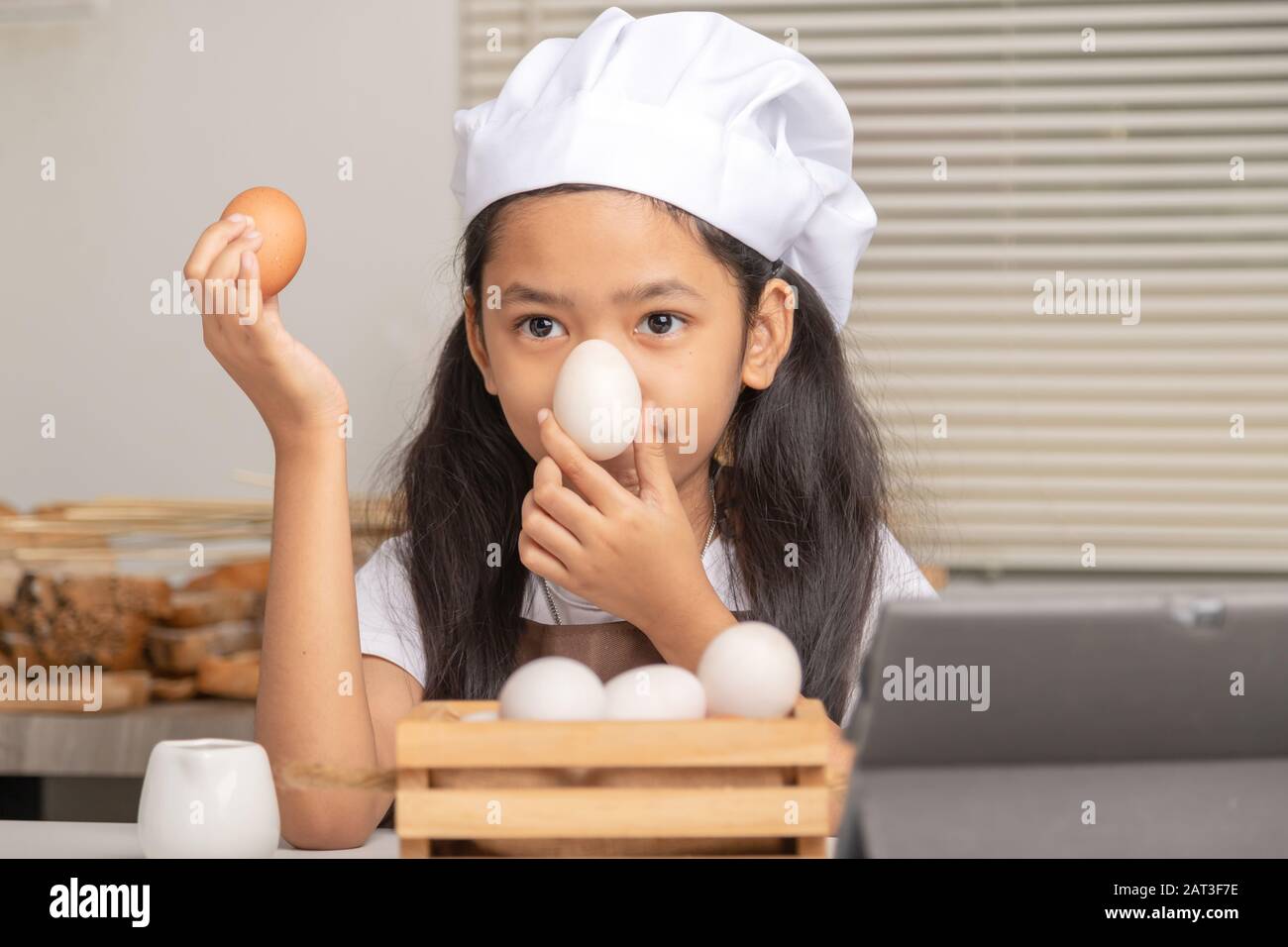 A little Asian girl wearing a white chef hat holding eggs and select a duck egg placed on her nose on a white cooking table in the kitchen. Stock Photo