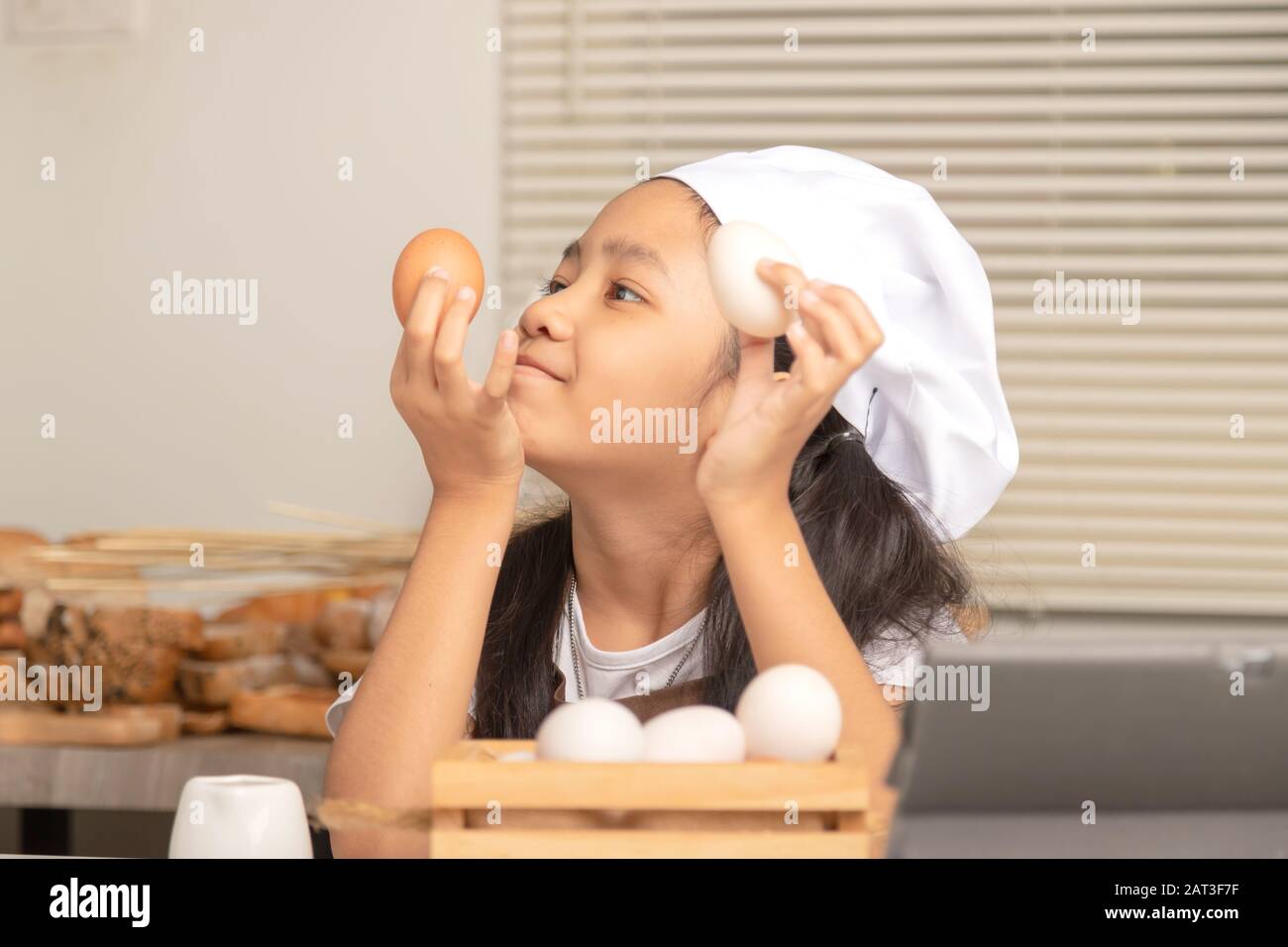 A little Asian girl wearing a white chef hat is holding and choosing between a duck egg and a chicken egg to make a bakery on a white cooking table in Stock Photo