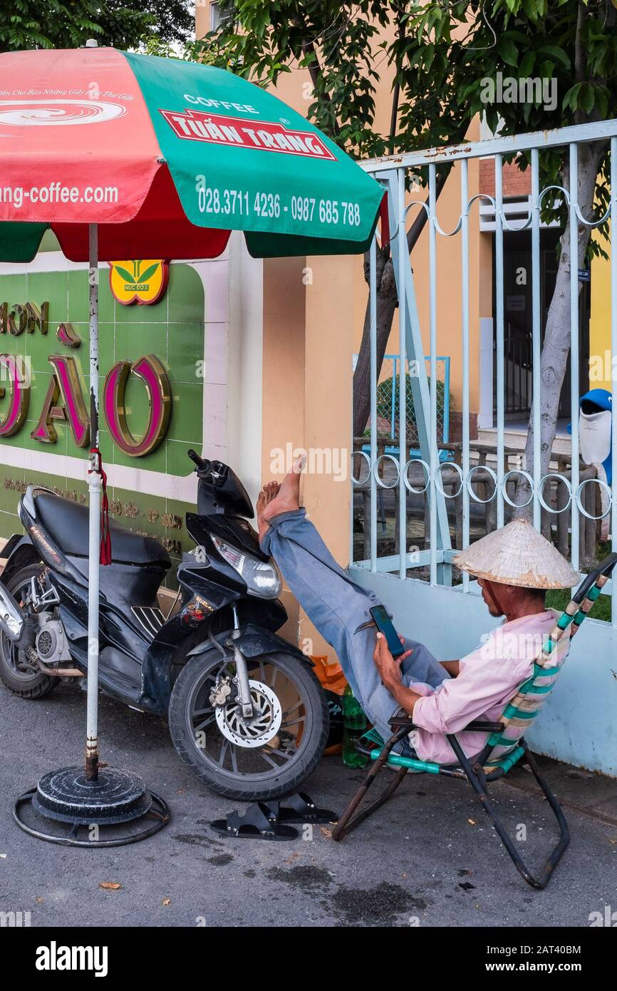 Man sat in chair looking at smart phone with his feet up on motorbike, Ho Chi Minh City, Vietnam Stock Photo