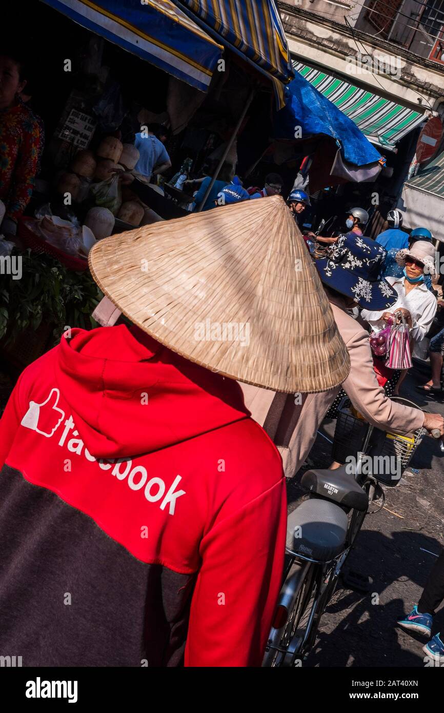 Man wearing Facebook top & conical hat walking through street market, Ho Chi Minh City, Vietnam Stock Photo