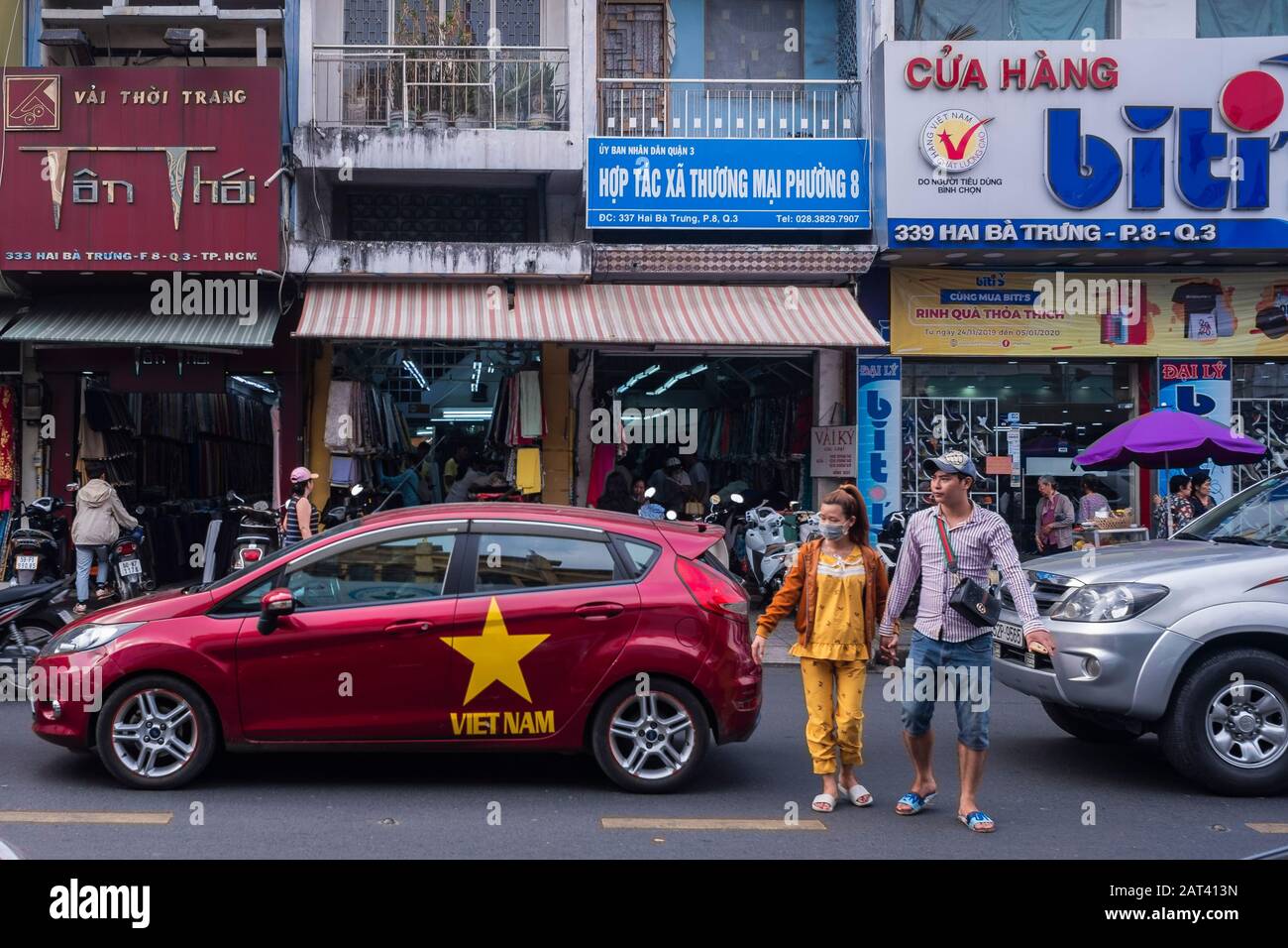 Couple crossing the road next to a car painted with the flag of Vietnam, Ho Chi Minh City, Vietnam Stock Photo