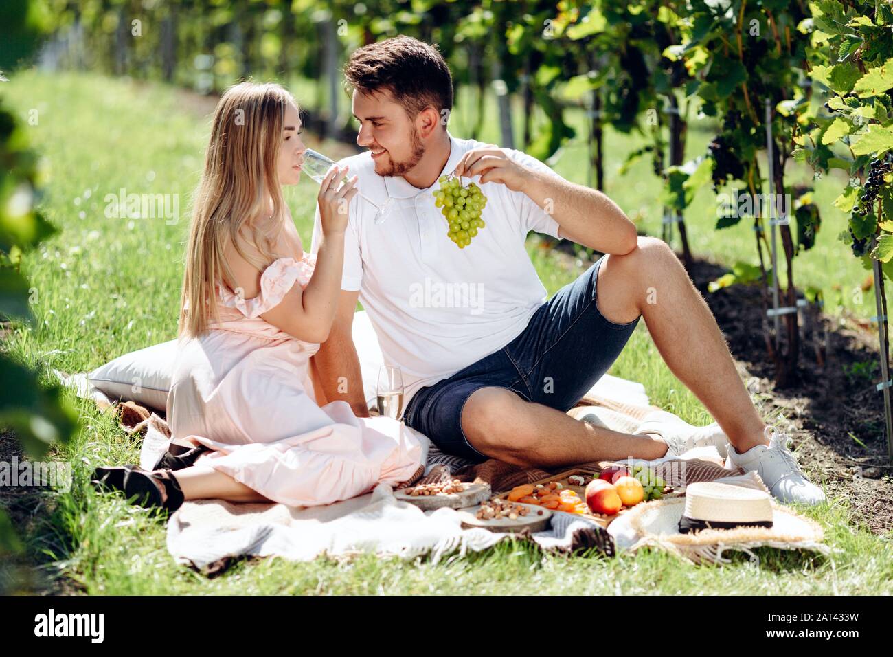 Enamored girl and boy sitting on blanket eating grapes and drinking wine during picnic in grape garden Stock Photo