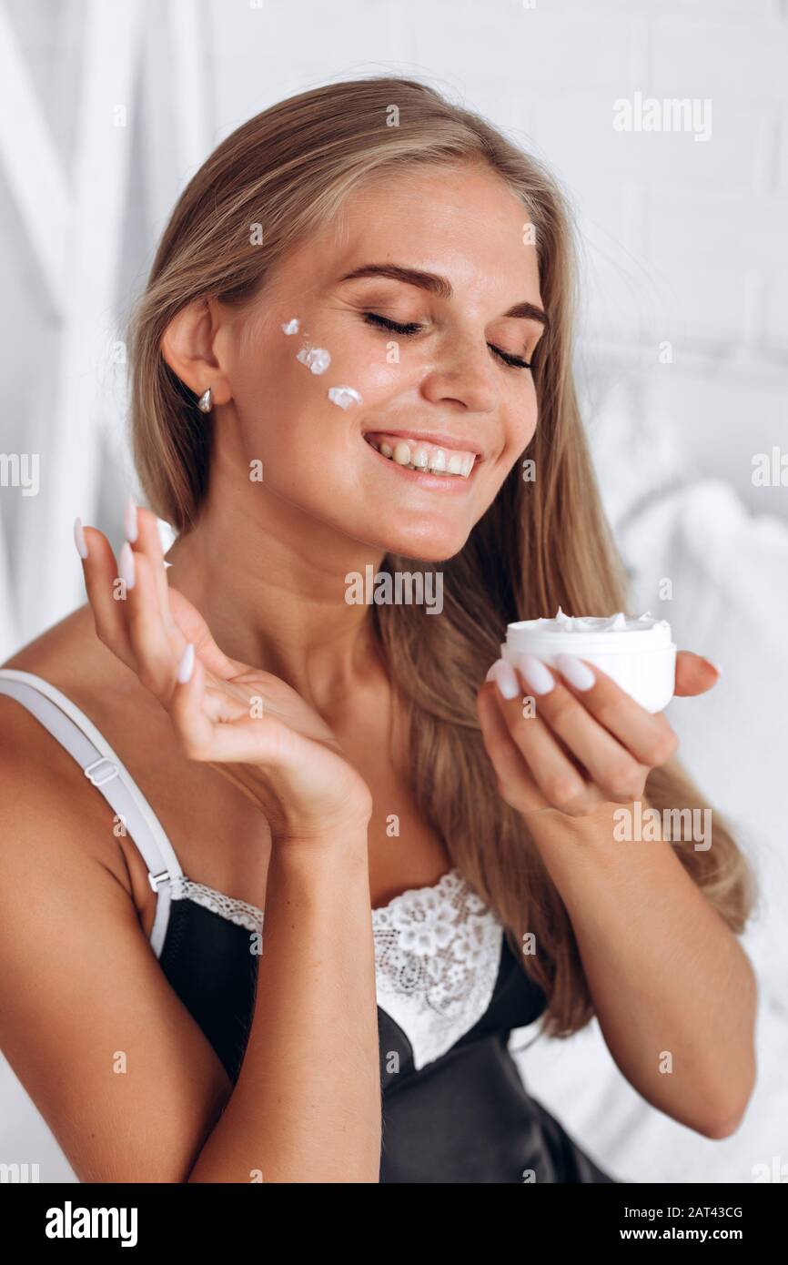 Smiling woman in black pajamas applies cream on face Stock Photo