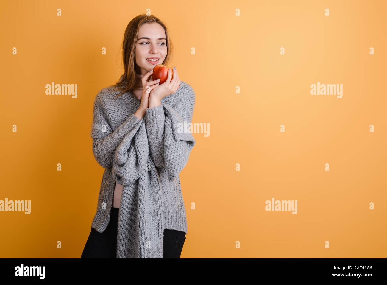 Girl in a warm sweater holds an apple and looks away. On a yellow background. Stock Photo