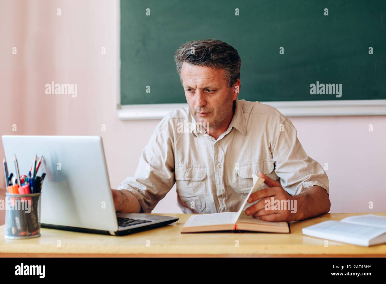 Middle age teacher sitting with open textbook and laptop and working.- Image Stock Photo