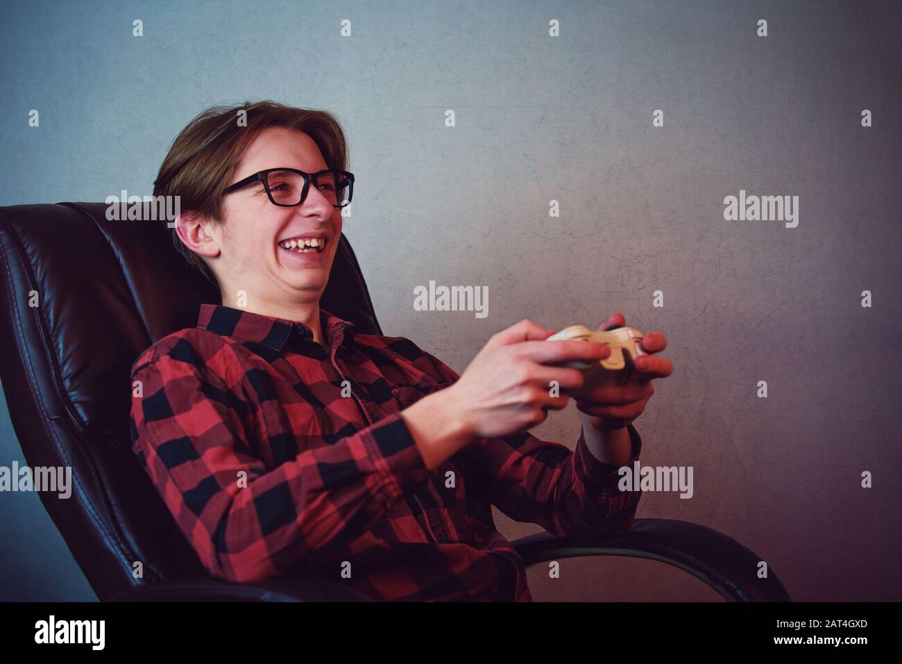 Happy teenage boy playing video games seated in his armchair. Excited adolescent wearing eyeglasses, holding joystick console smiling to camera, try t Stock Photo