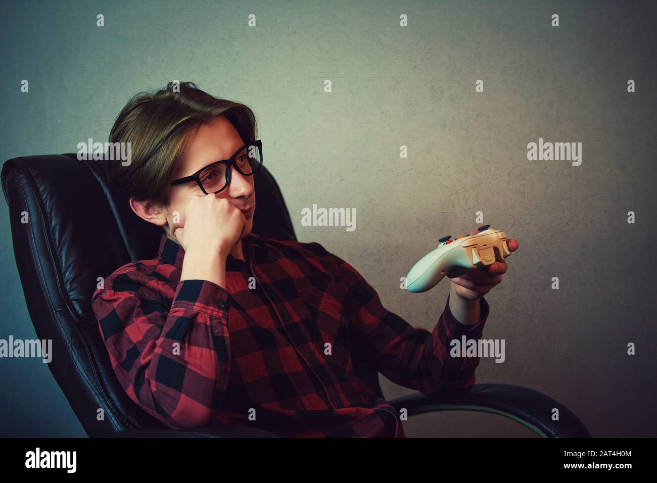 Bored adolescent boy seated in his armchair late night playing video games inside a dark room background. Sleepy boy holding joystick console, being i Stock Photo