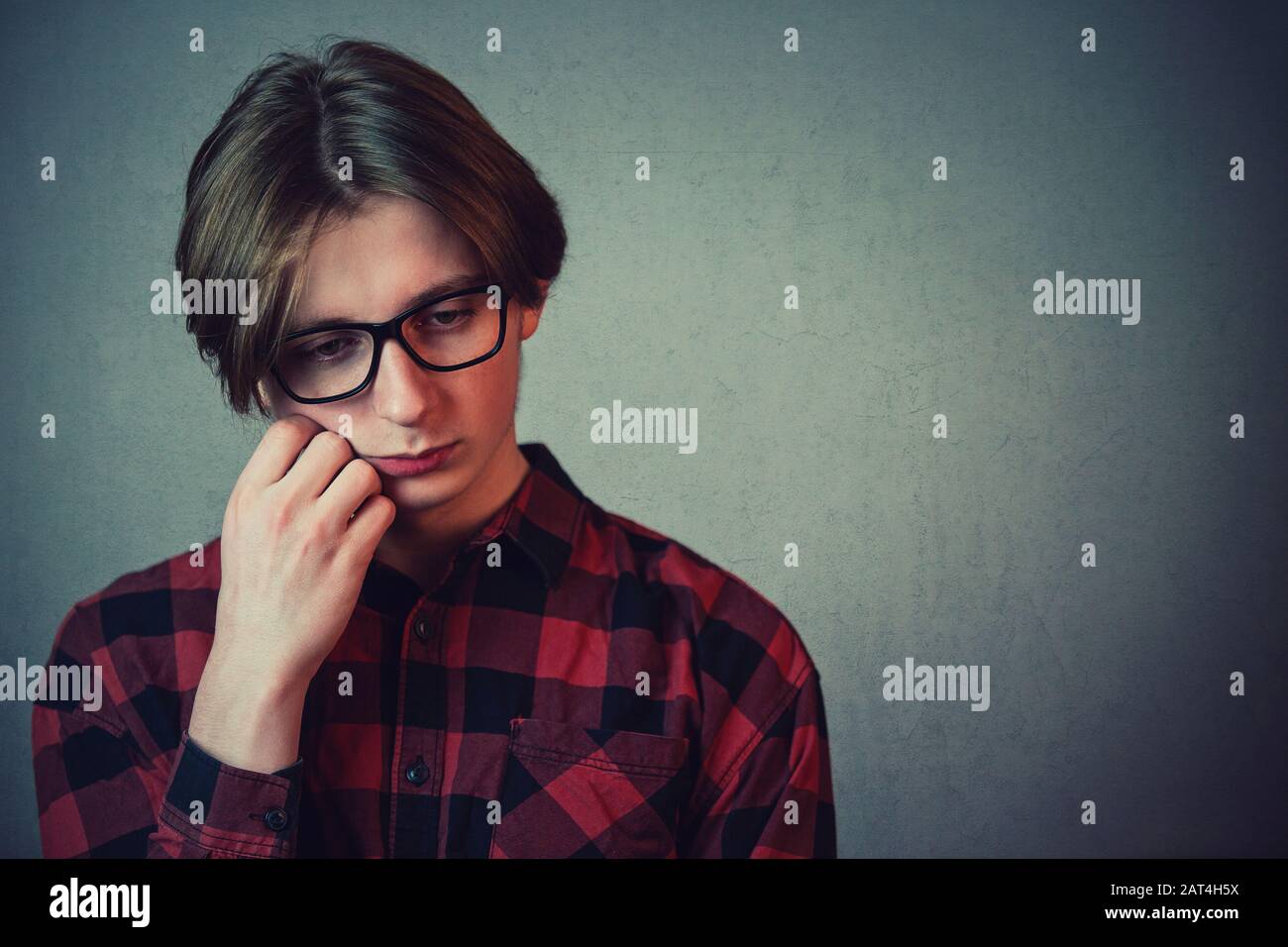 Closeup portrait of sad and thoughtful teen guy, hand under cheek, looking down as feel guilty and ashamed isolated on grey wall. Upset boy experienci Stock Photo