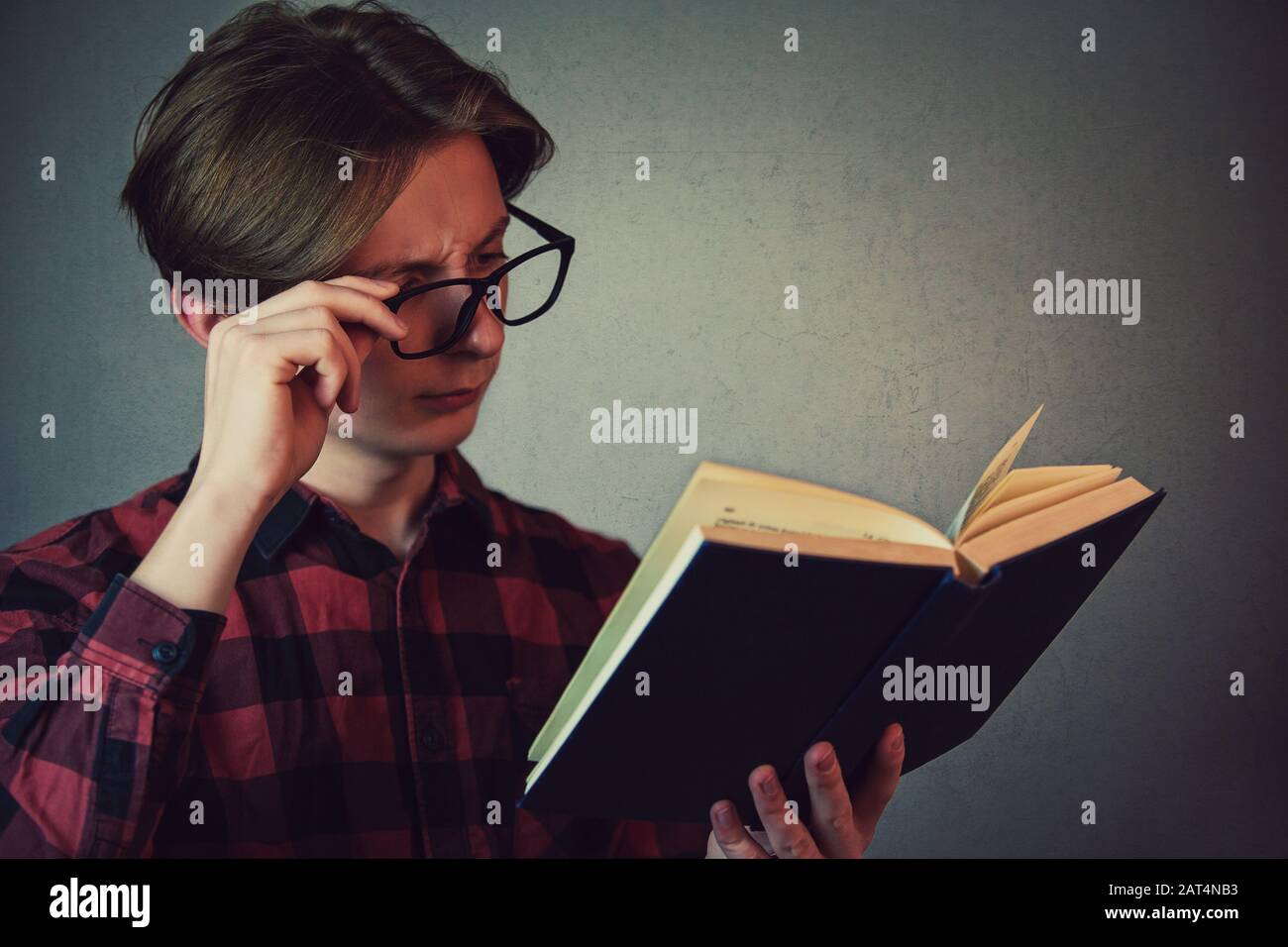 Young man student, fixing glasses, trying to read a book, poor sight farsightedness, myopia. Teenage reader eyes problem, suffering eyestrain, sore an Stock Photo