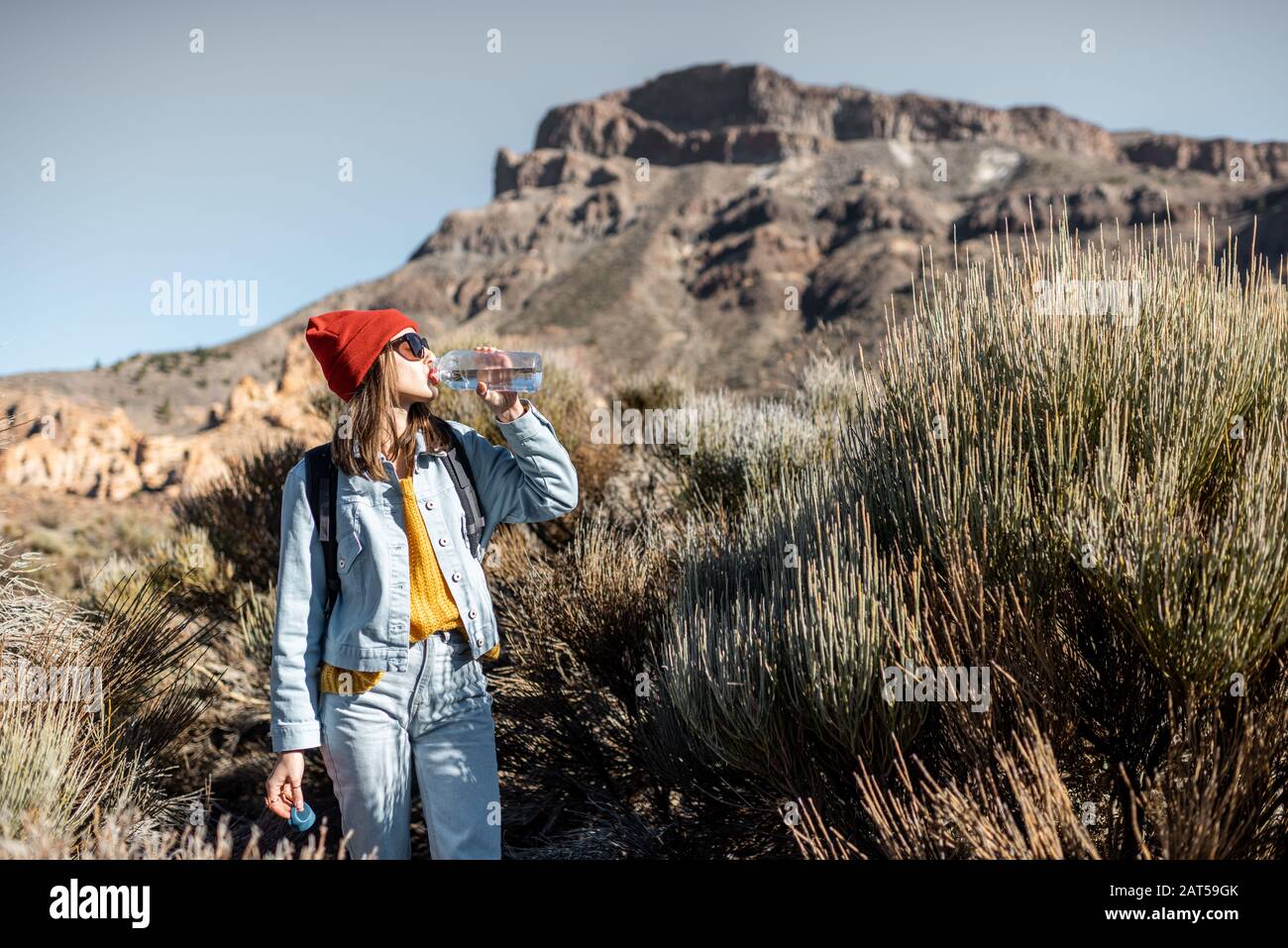Young woman in red hat traveling with backpack on the volcano valley, feeling thirst and drinking water Stock Photo