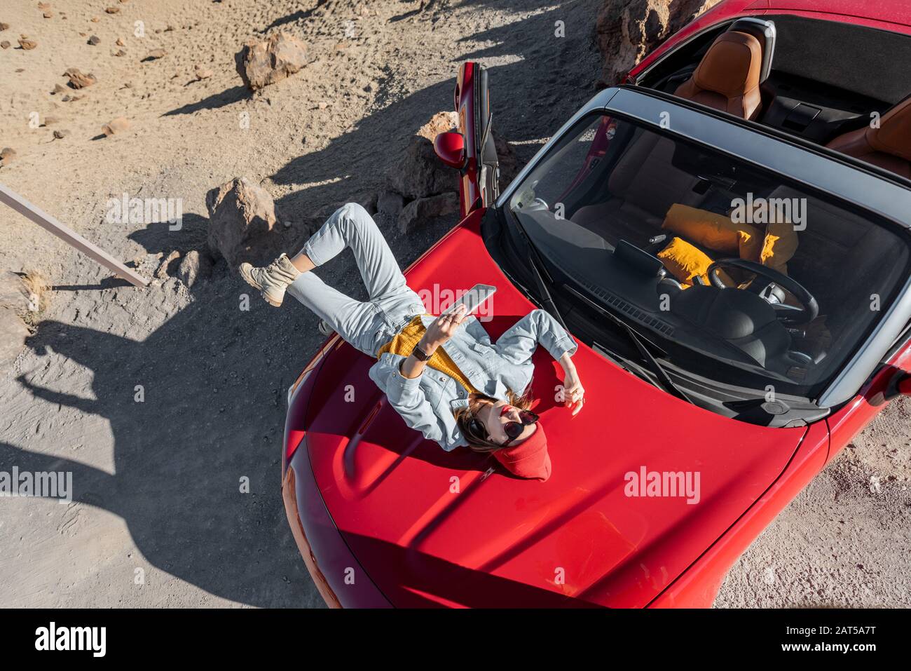 Lifestyle portrait of a young woman enjoying road trip on the desert valley, lying on the car hood and photographing on phone. View from above Stock Photo