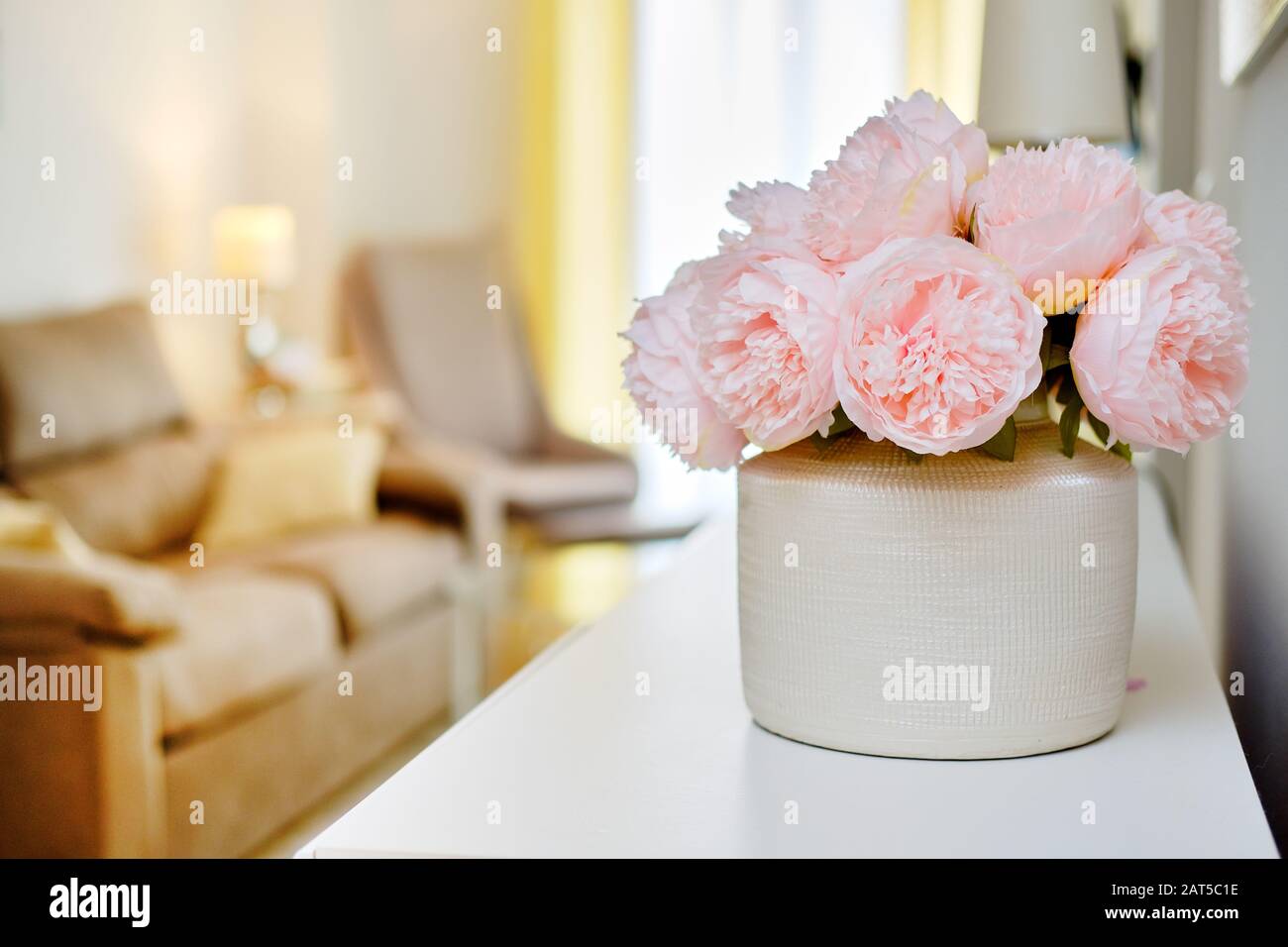 Pretty pink colour peonies flowers in white vase inside of light warm living room, selective close up focus on bouquet, no people Stock Photo