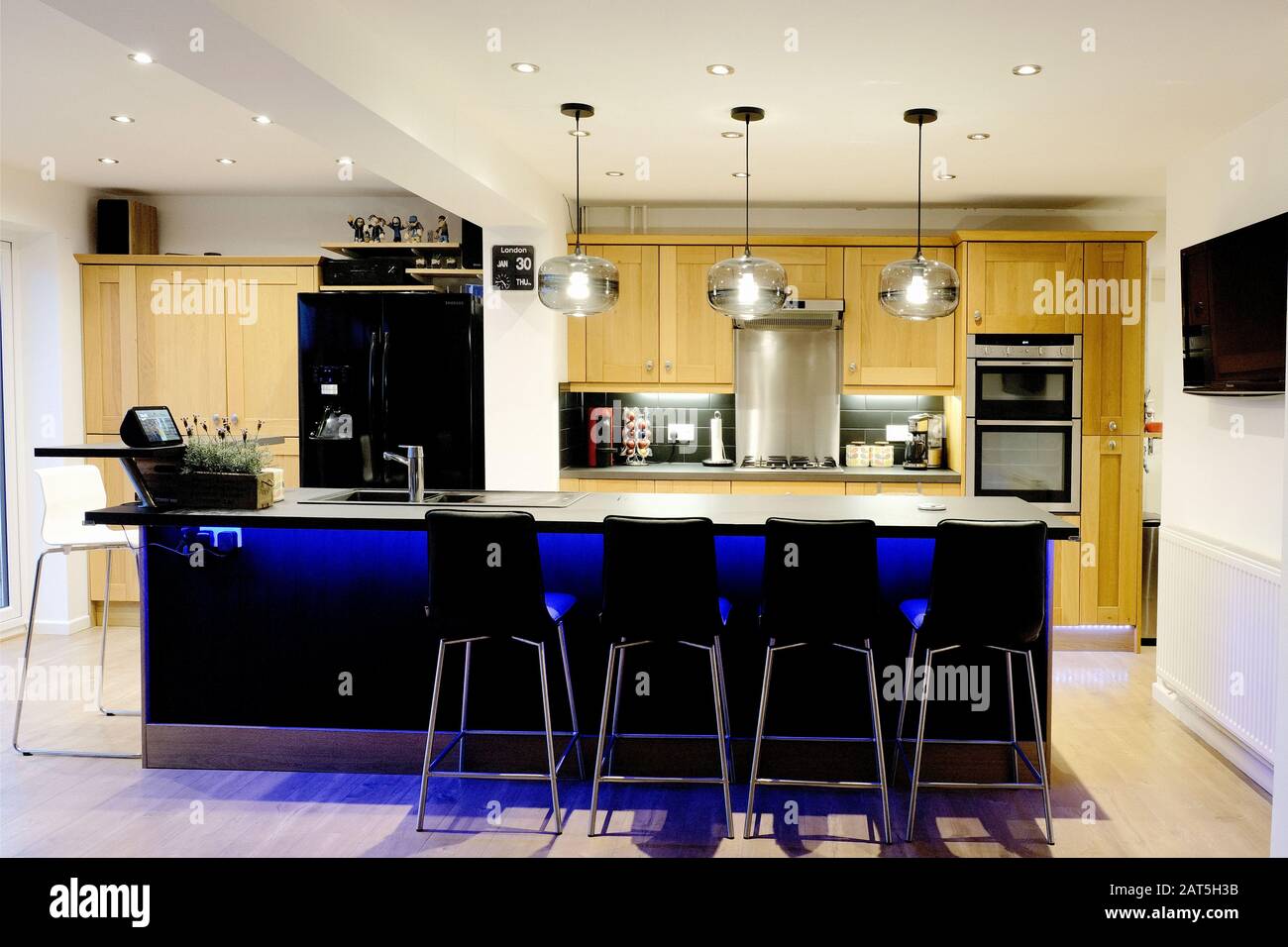A smart modern kitchen in a modern, British, home featuring a large island with bar stools and wooden kitchen units. The kitchen is illuminated. Stock Photo