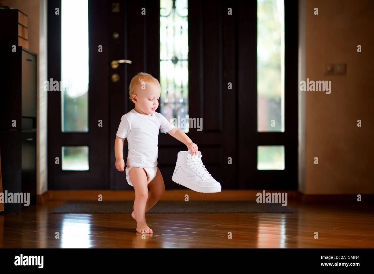 little girl in a white T-shirt and diaper holds a white sneaker standing in the hallway at home Stock Photo