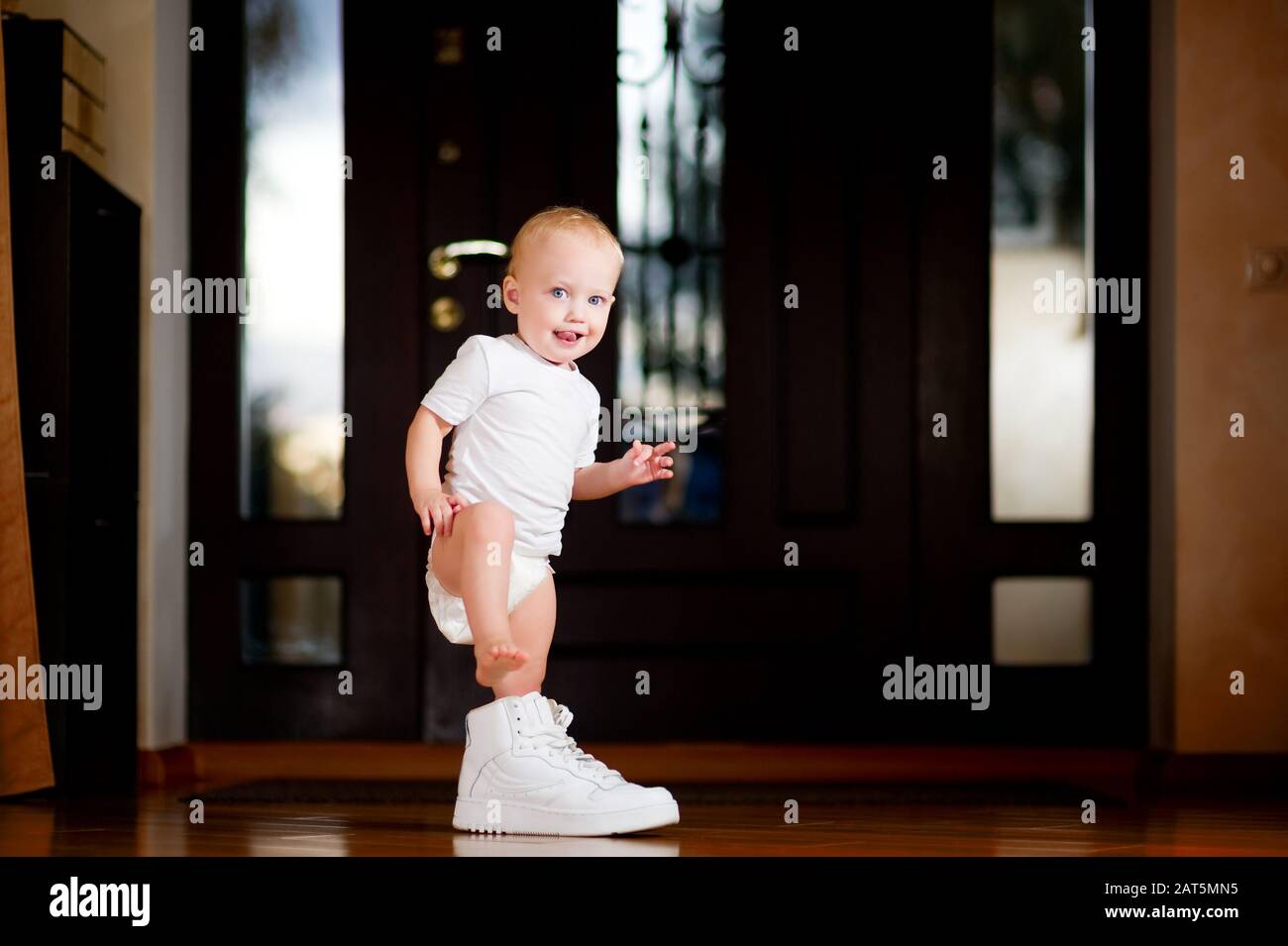 baby girl trying to put on a big white sneaker of an adult standing indoors Stock Photo