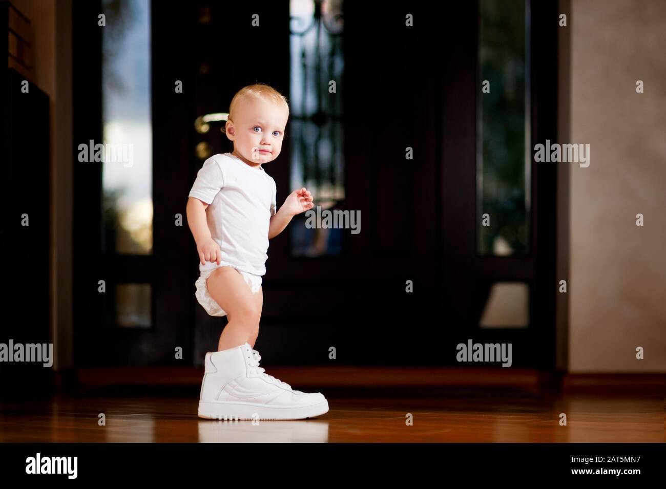 child put on adult white sneaker while standing in hallway Stock Photo