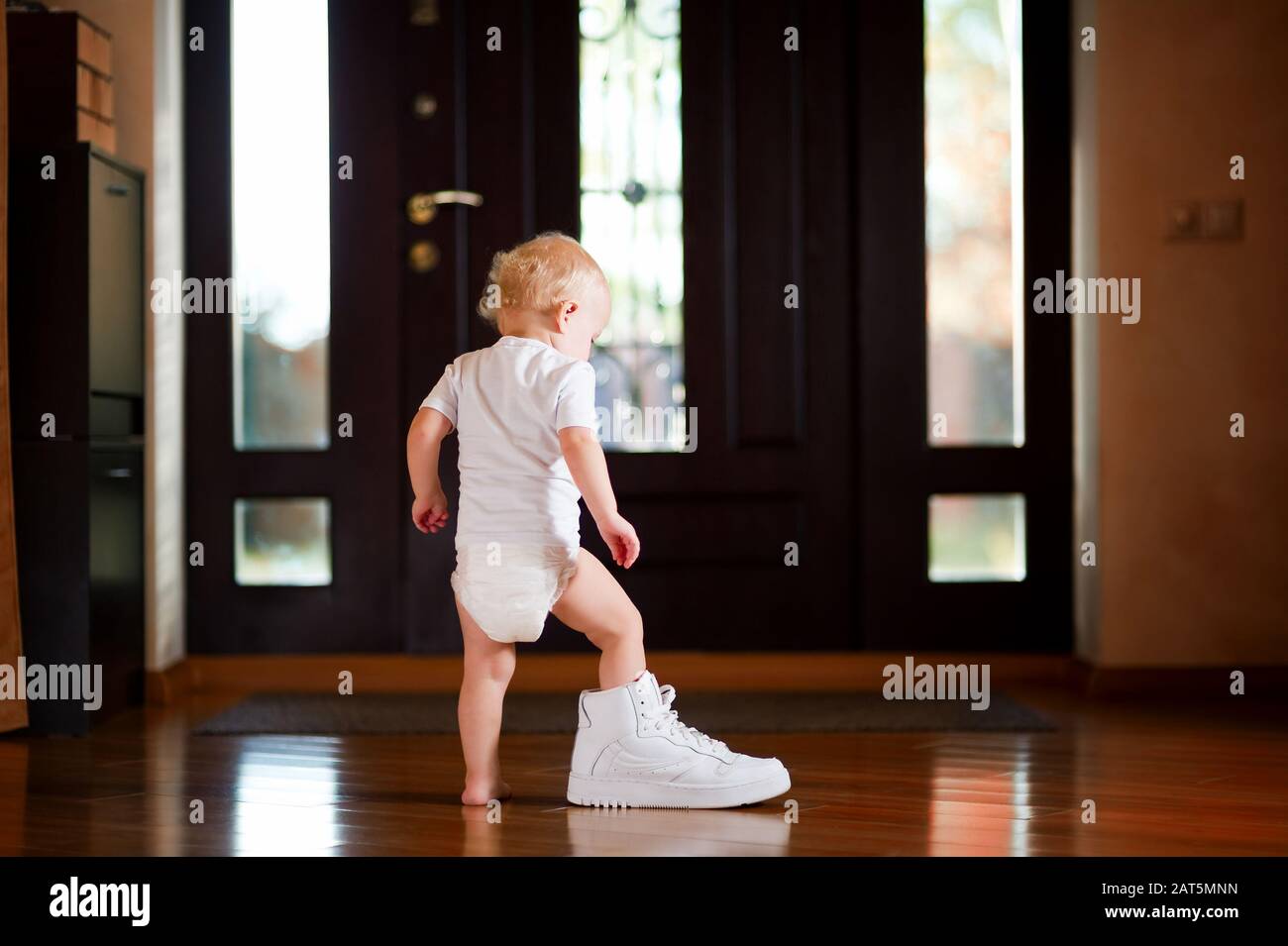 baby girl in a white T-shirt and diaper puts on a white dad's sneaker, standing in the hallway at home Stock Photo