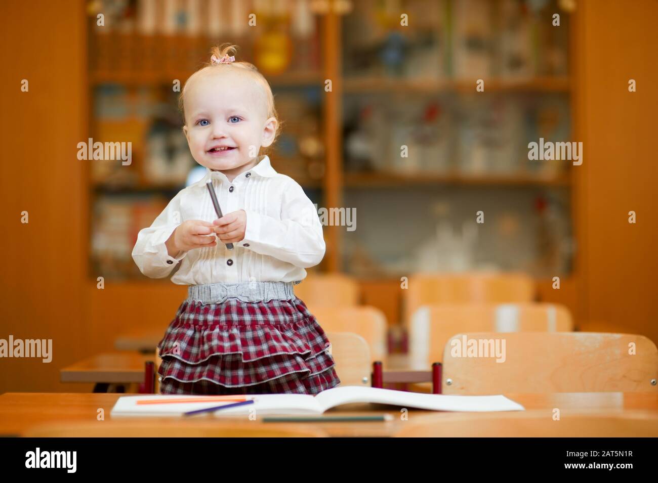 Baby Girl Stands On A Chair At A School Desk And Smile Stock Photo