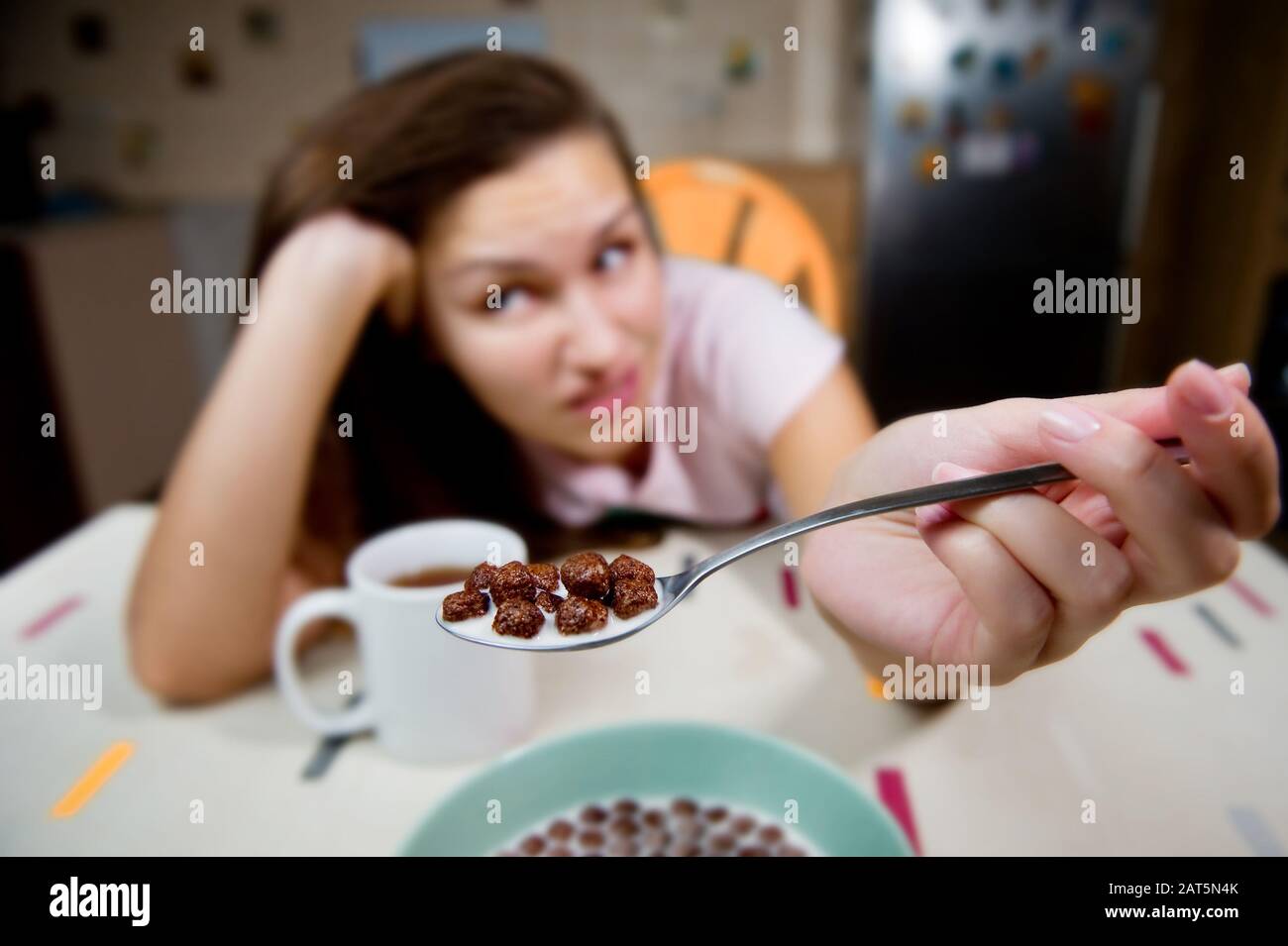 A young woman shows a spoon with chocolate balls and expresses a reluctance to eat them. Chocolate cereal with milk in focus and girl with grimace in Stock Photo