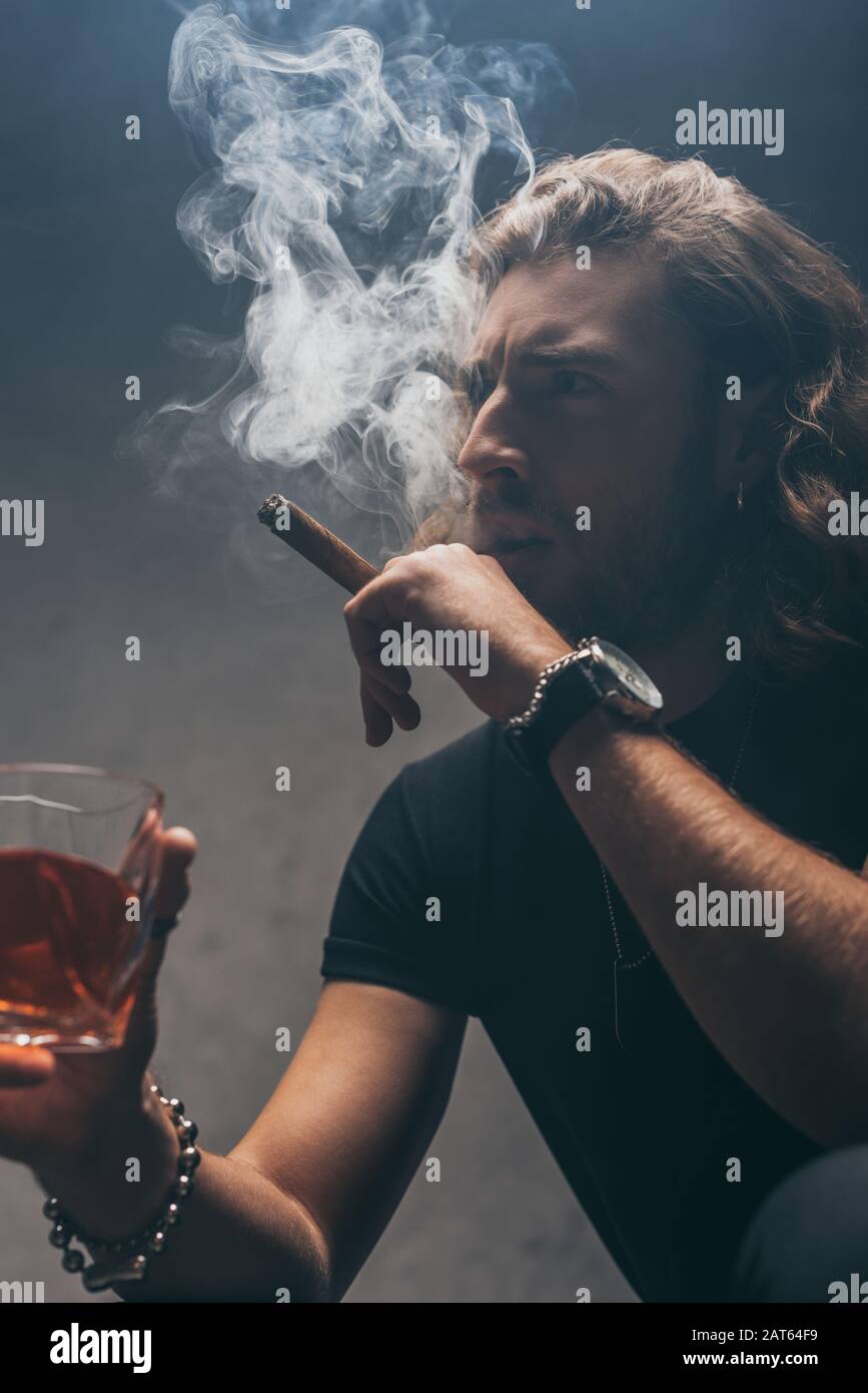 tense fashionable businessman in black outfit smoking cigar and drinking whiskey near grey wall Stock Photo