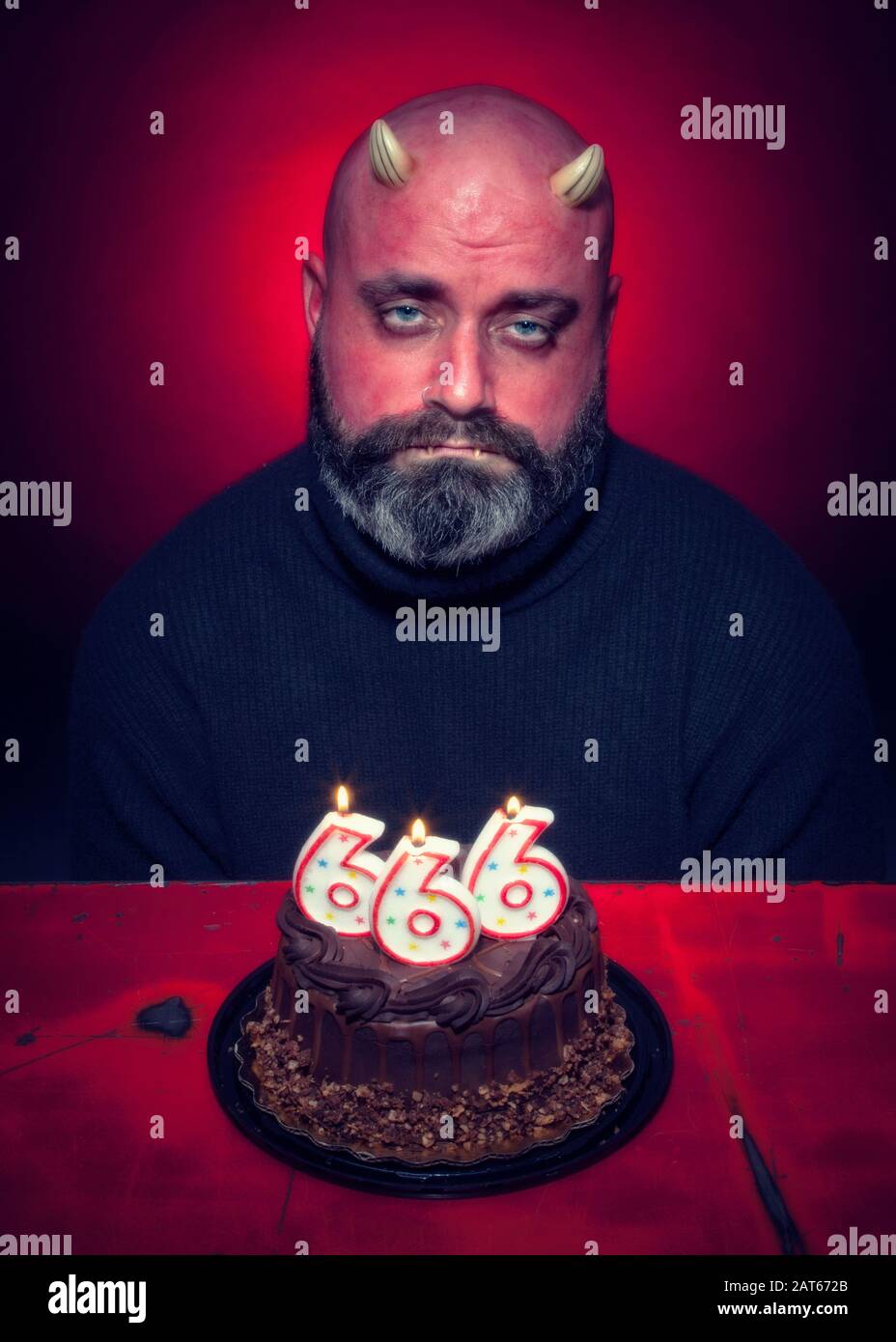 A sad man sits in front of a devil's food chocolate birthday cake with number 666 candles with a glowing red background Stock Photo