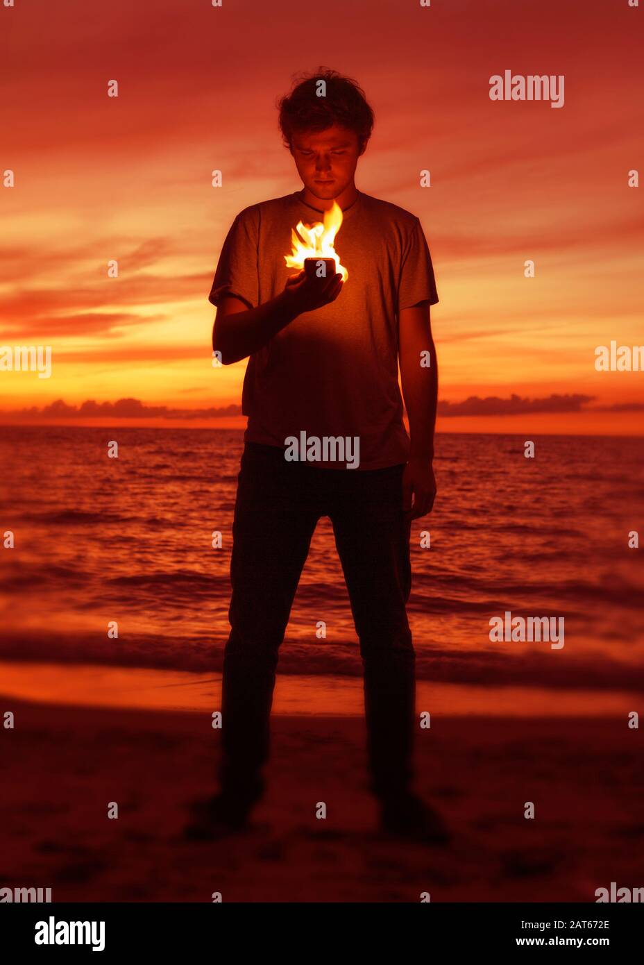 A young man stands on a beach at sunset and looks at his burning smart phone as flames illuminate his face Stock Photo