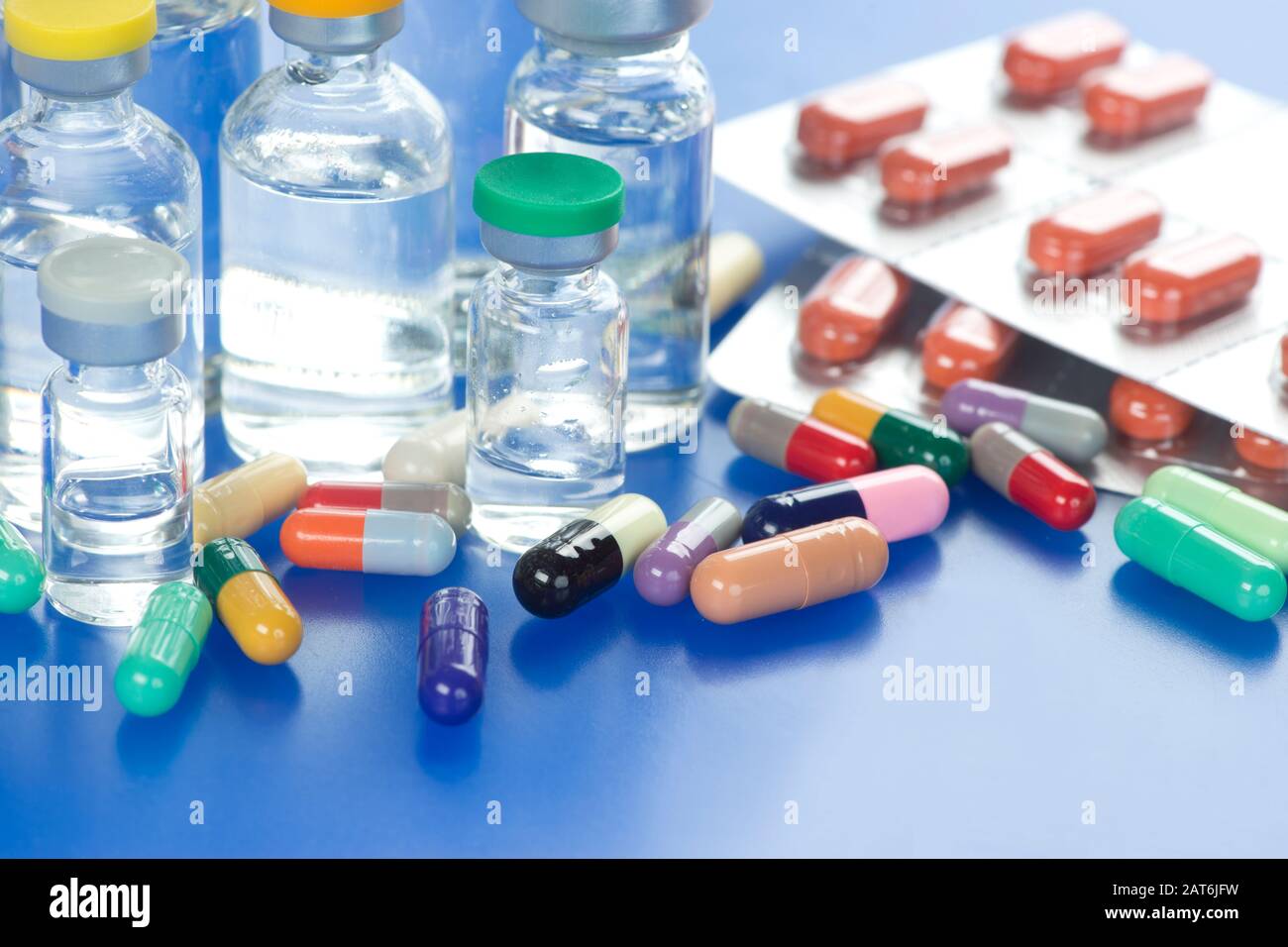Many different colored medication capsules and foil pack with various injection vaccine vials on blue colored tray. Stock Photo