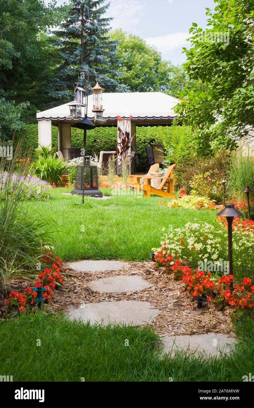 Grey flagstone footpath through mulch borders planted with Miscanthus - Ornamental grass plants, red Begonia flowers and gazebo in backyard garden Stock Photo
