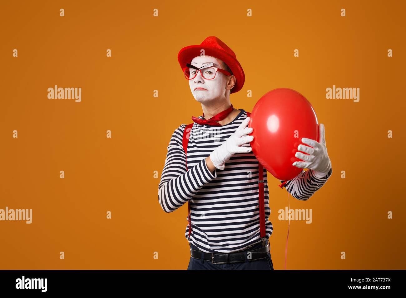 Sad clown in vest and red hat with balloon in his hand on orange empty background Stock Photo