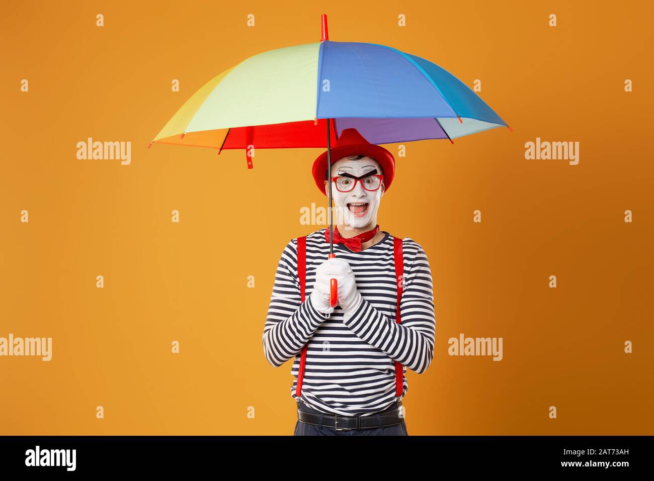 Funny mime with multi-colored umbrella on empty orange background in studio Stock Photo